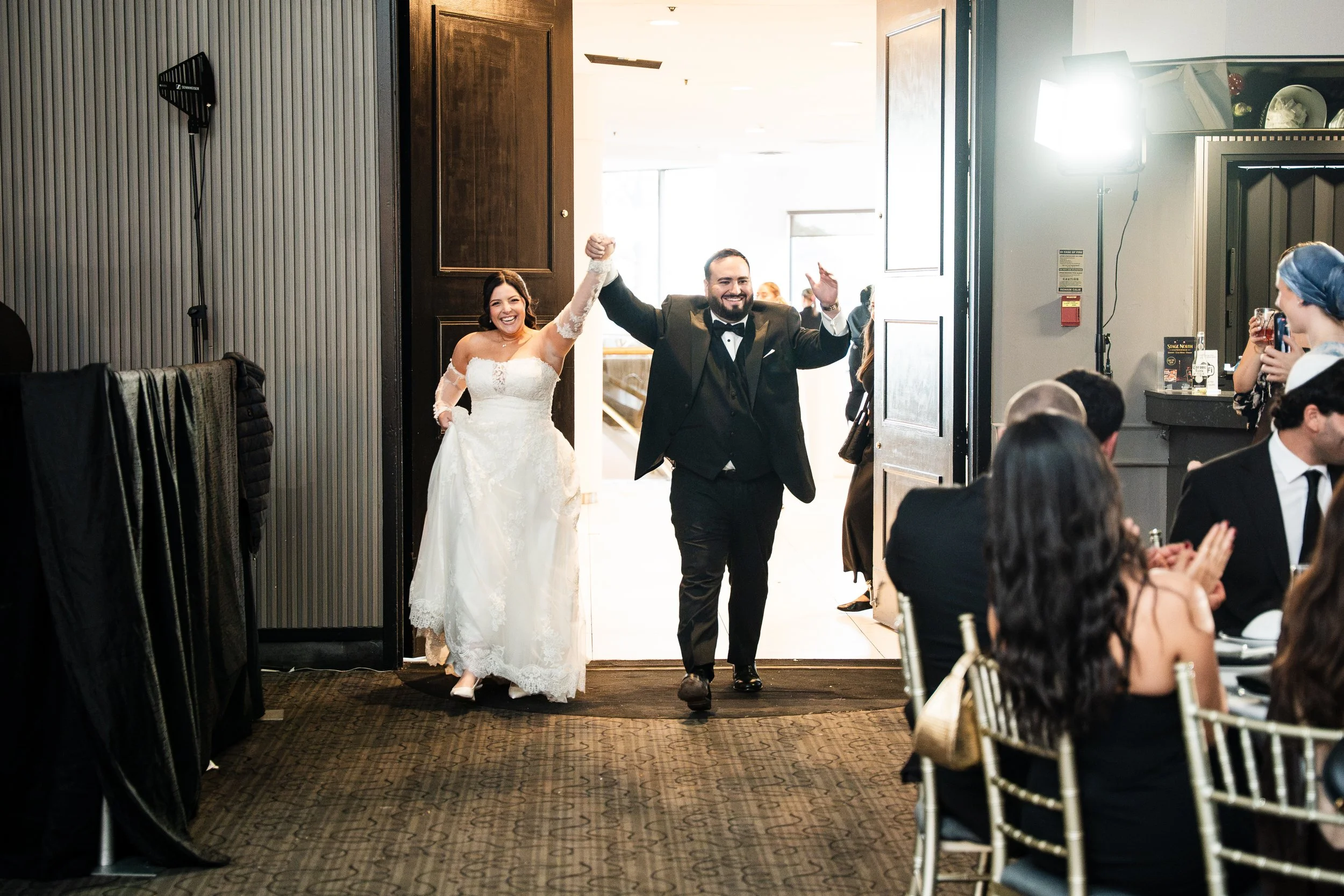A newlywed couple walks through a doorway into a celebration hall, smiling and raising their arms in happiness. The bride wears a white lace wedding dress, and the groom is dressed in a black tuxedo. Guests are seated on either side, some clapping an