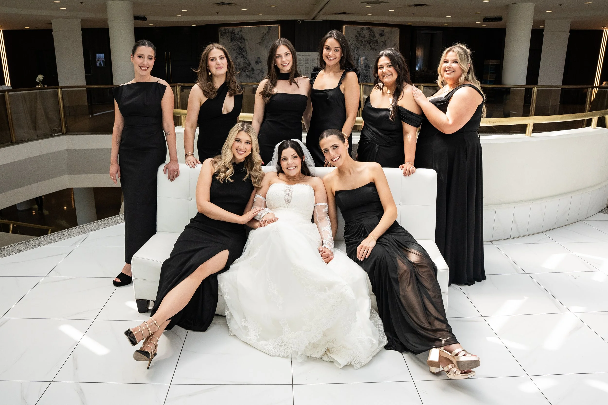 A group of women in black dresses and a bride in a white gown sitting on a white sofa in a modern, indoor setting with white tile floors and a curved railing behind them.