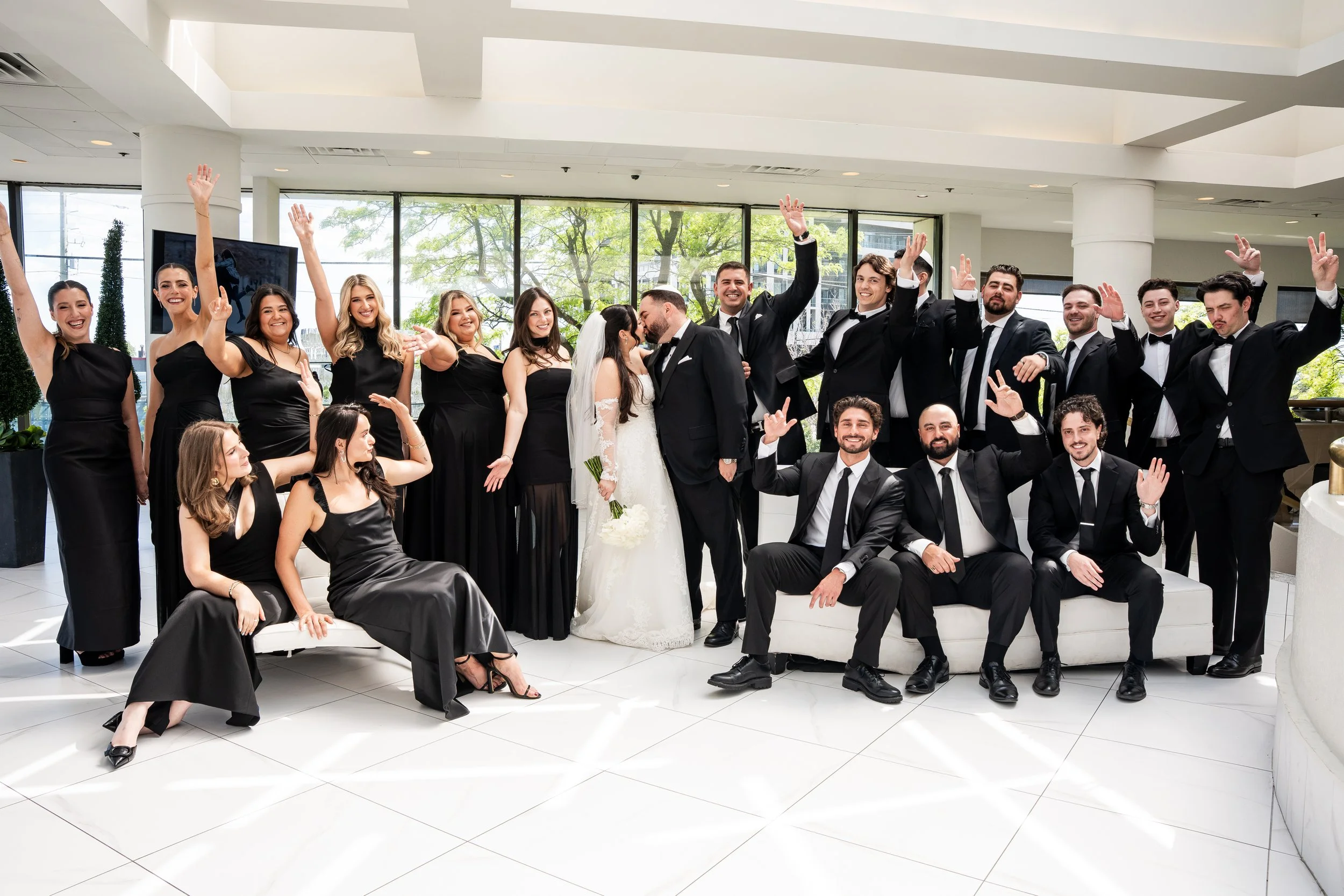 Group of people dressed in formal attire celebrating at a wedding, with some raising their hands and others sitting or standing in a bright, modern indoor space with large windows and green trees visible outside.