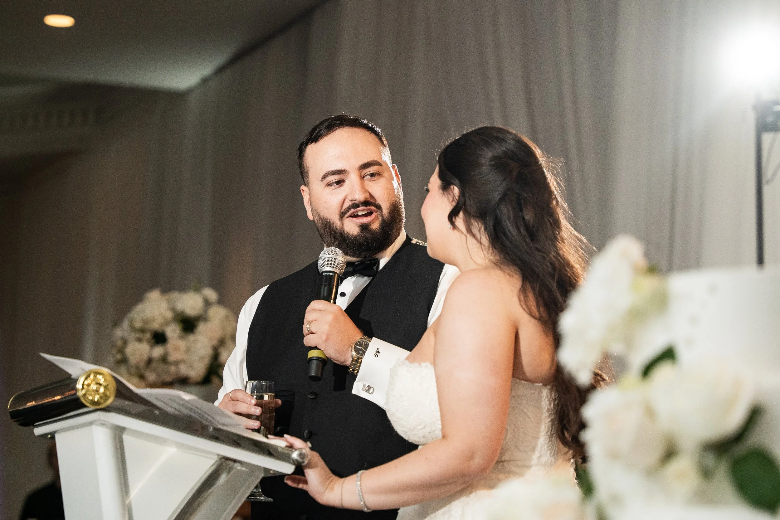 A groom and bride exchanging vows at their wedding ceremony. The groom is holding a microphone and a glass, wearing a tuxedo, and speaking to the bride who is dressed in a wedding gown.