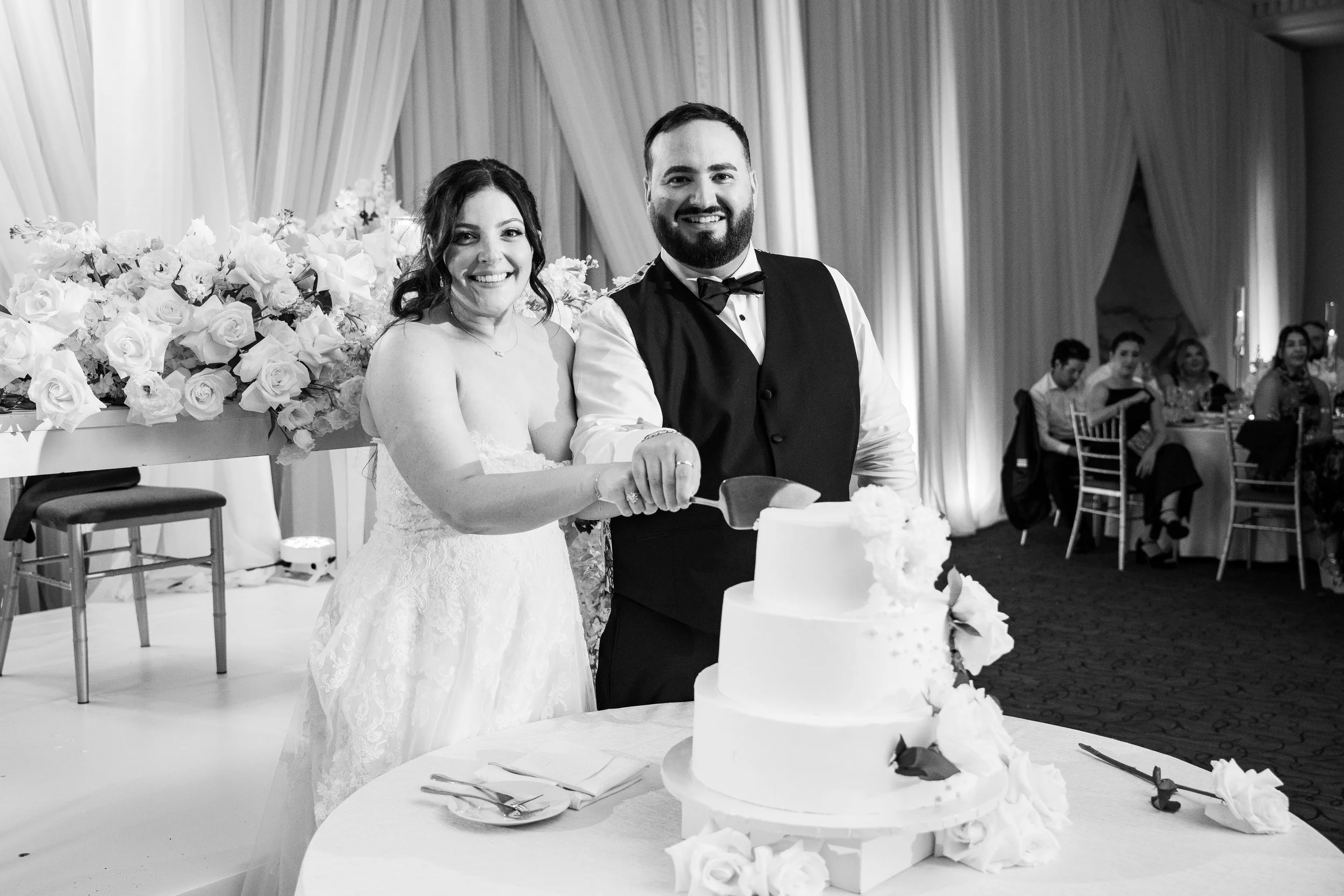 Black and white photo of a newlywed couple cutting their wedding cake at a reception. The bride is wearing a wedding dress and has long dark hair, and the groom is in a tuxedo with a bow tie. There are guests seated at tables in the background, and a