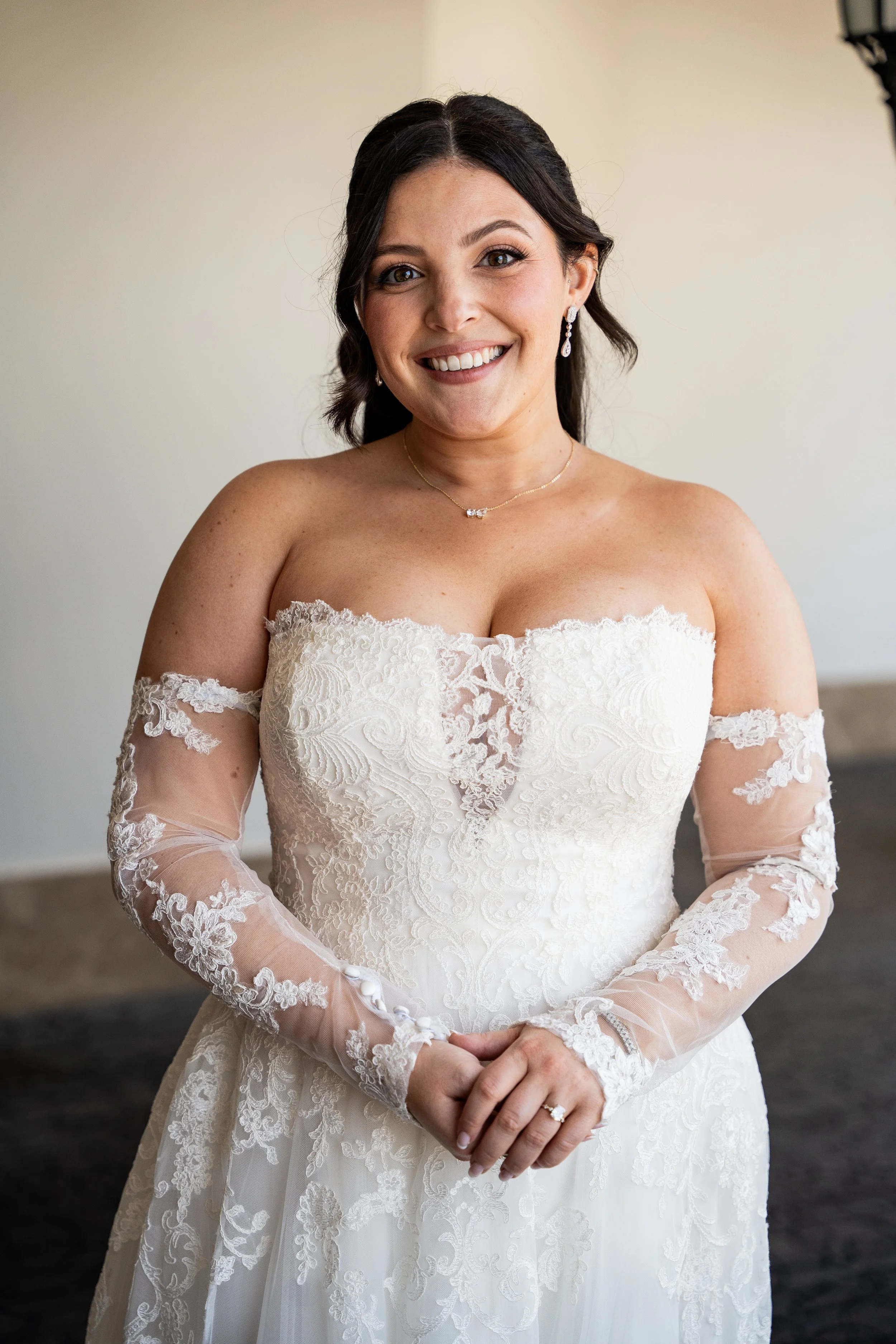 A woman in a white lace wedding dress with sheer lace long sleeves, wearing jewelry, smiling with hands clasped in front of her, standing indoors.