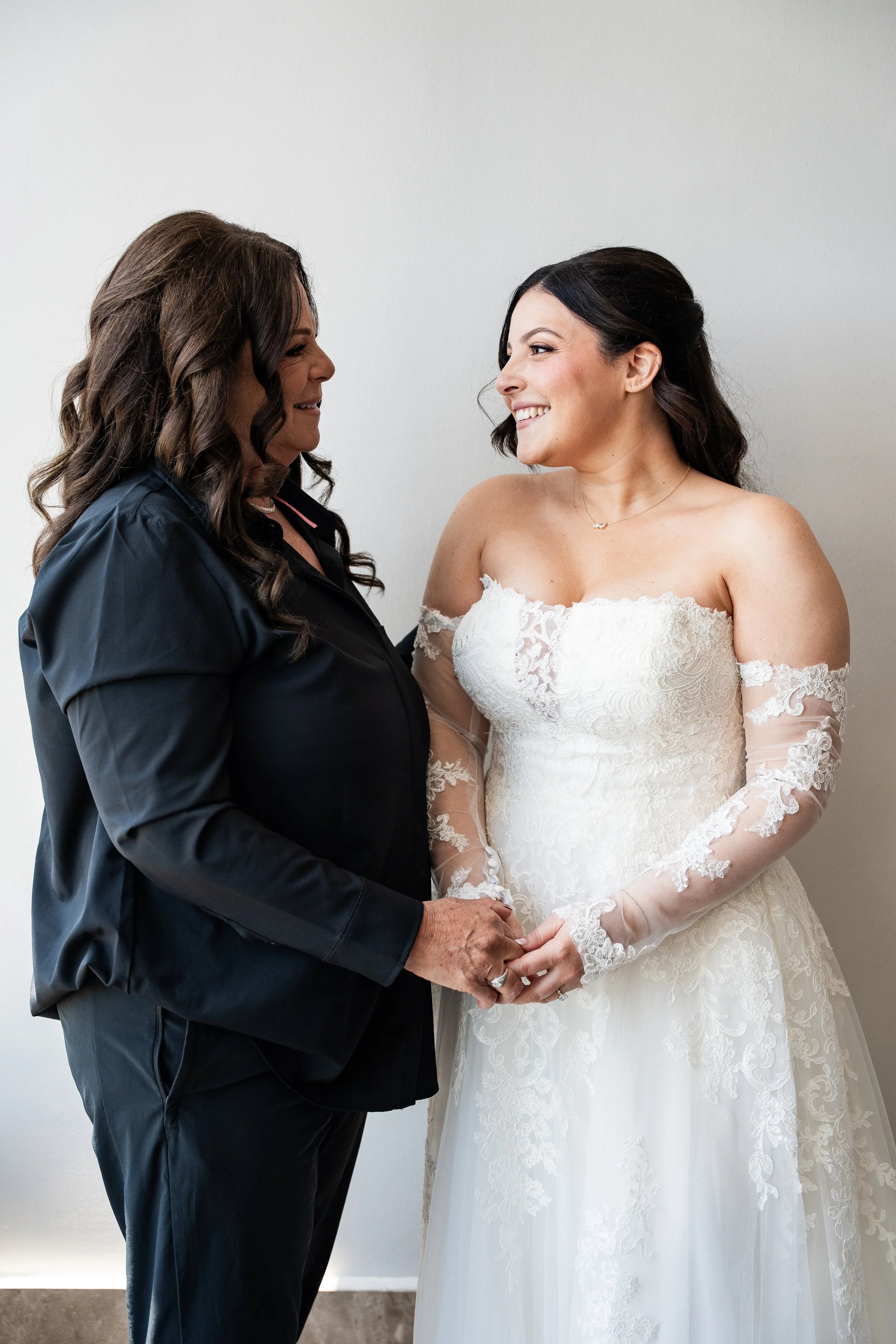 A bride and a woman holding hands and smiling at each other in a wedding preparation.