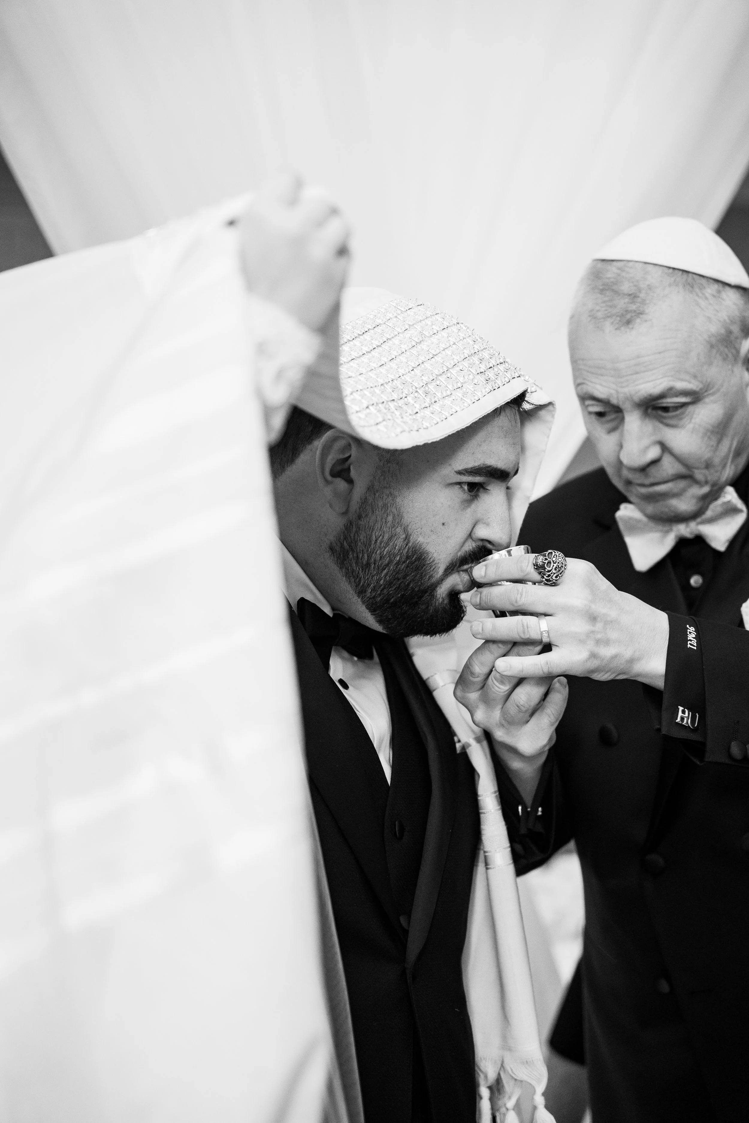 Two men, one with a beard and another with a shaved head, wearing traditional Jewish head coverings, are engaged in a religious ceremony involving drinking from a cup. The man with the shaved head is holding the cup, and the other man is guiding or a
