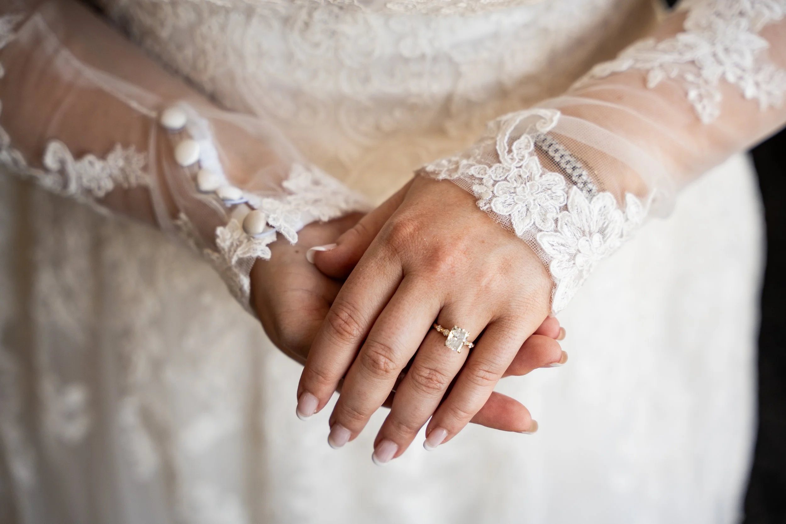 Close-up of two hands holding each other, one hand wearing a wedding ring with a large gemstone, both hands have neatly manicured fingernails. The background shows the sleeve of a lace wedding dress with intricate embroidery and buttons.