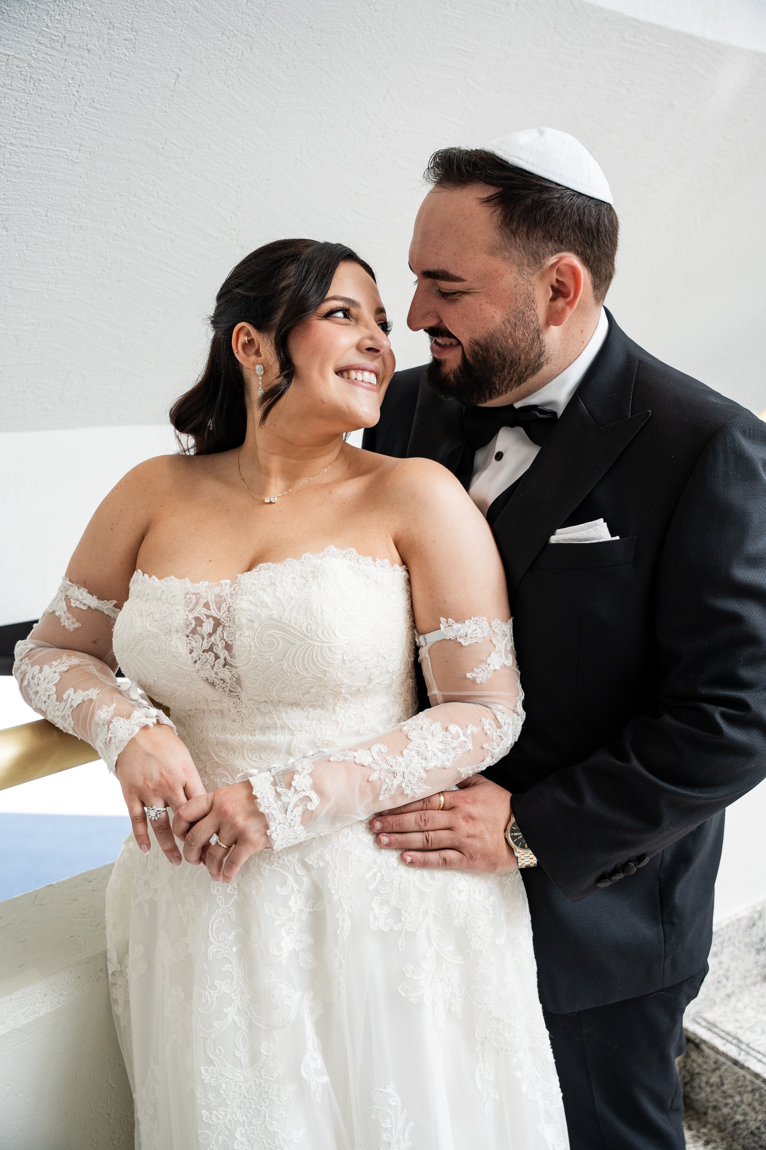 A bride and groom smiling at each other, with the bride wearing a lace wedding dress and the groom in a tuxedo and yarmulke, inside a plain room with white walls.