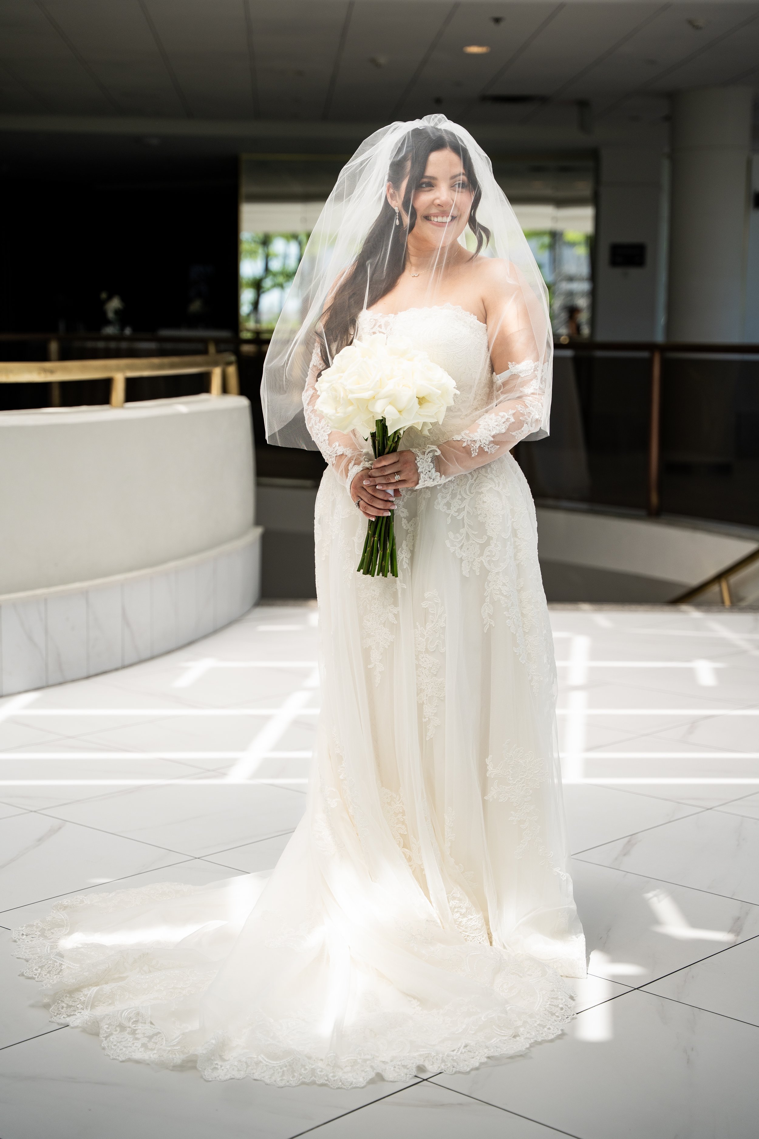 A bride in a white wedding dress holding a bouquet of white flowers inside a modern building with white tiled flooring and large windows.