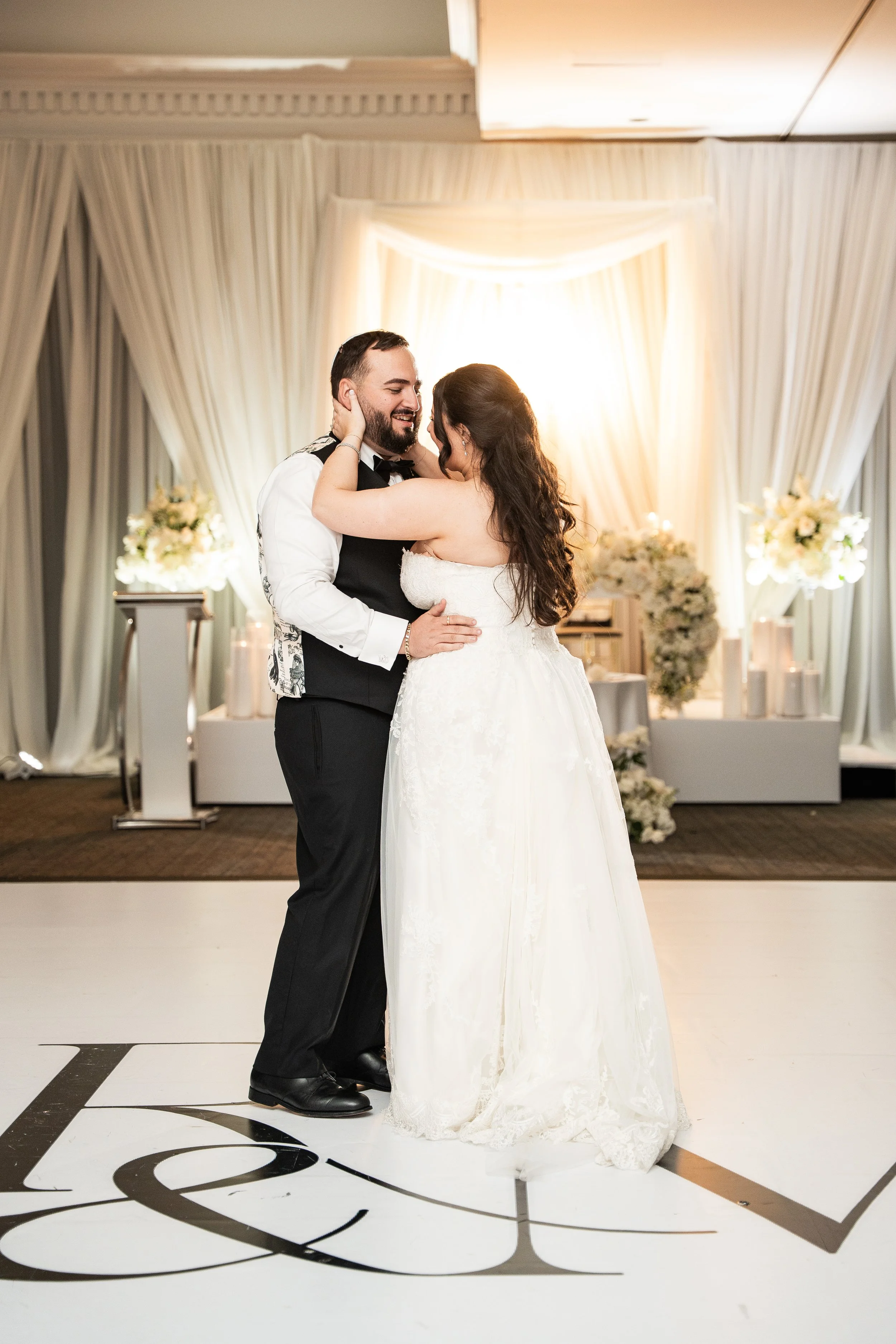 A bride and groom are dancing on a wedding dance floor, with the bride in a white wedding dress and the groom in a black vest, white shirt, and black bow tie, in a decorated venue with draped curtains and floral arrangements.