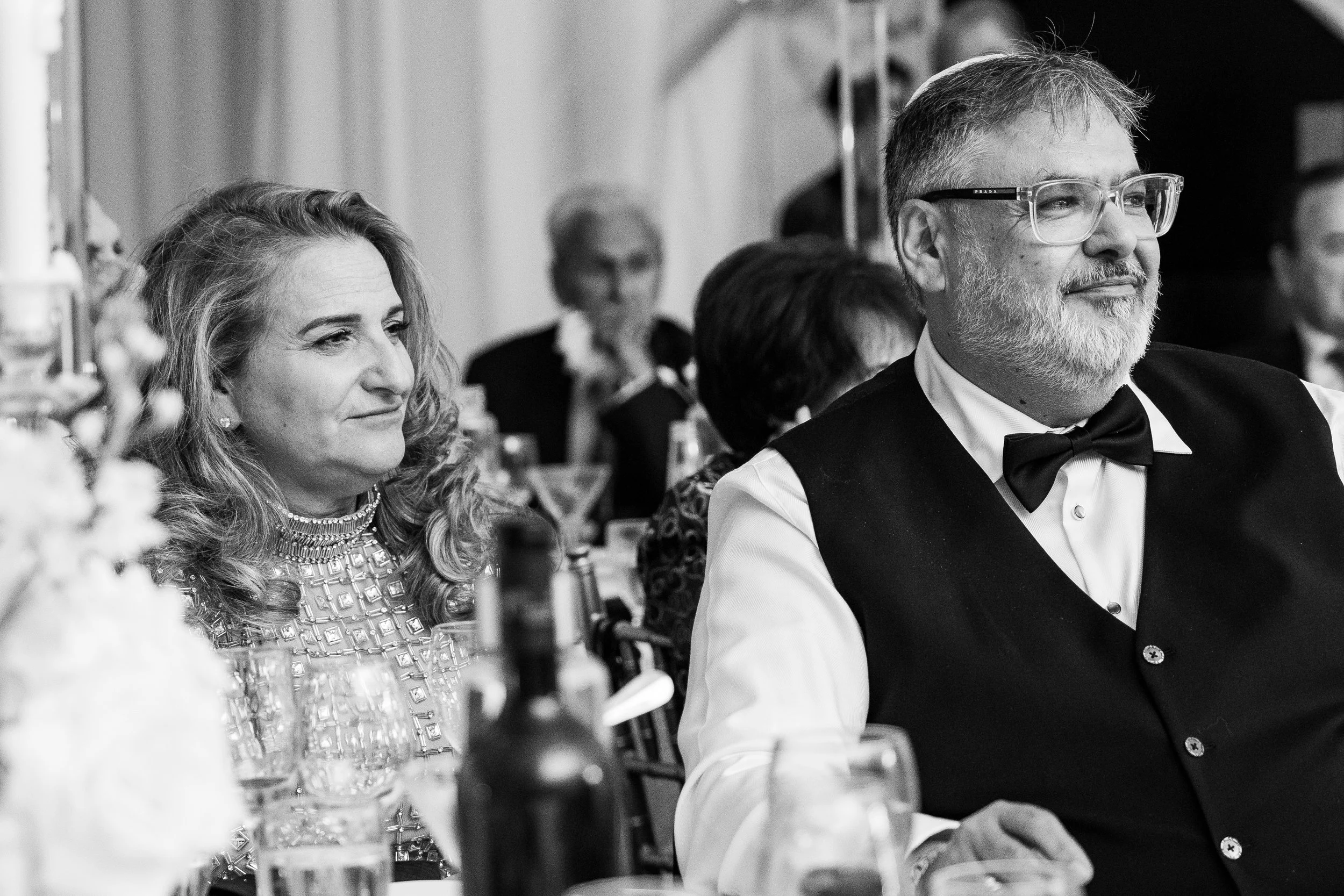 People at a formal event, including a woman with long wavy hair and a sparkling dress, and a man with glasses, a beard, wearing a tuxedo with a bowtie. They are seated at a table with wine glasses and bottled water, with other guests visible in the b