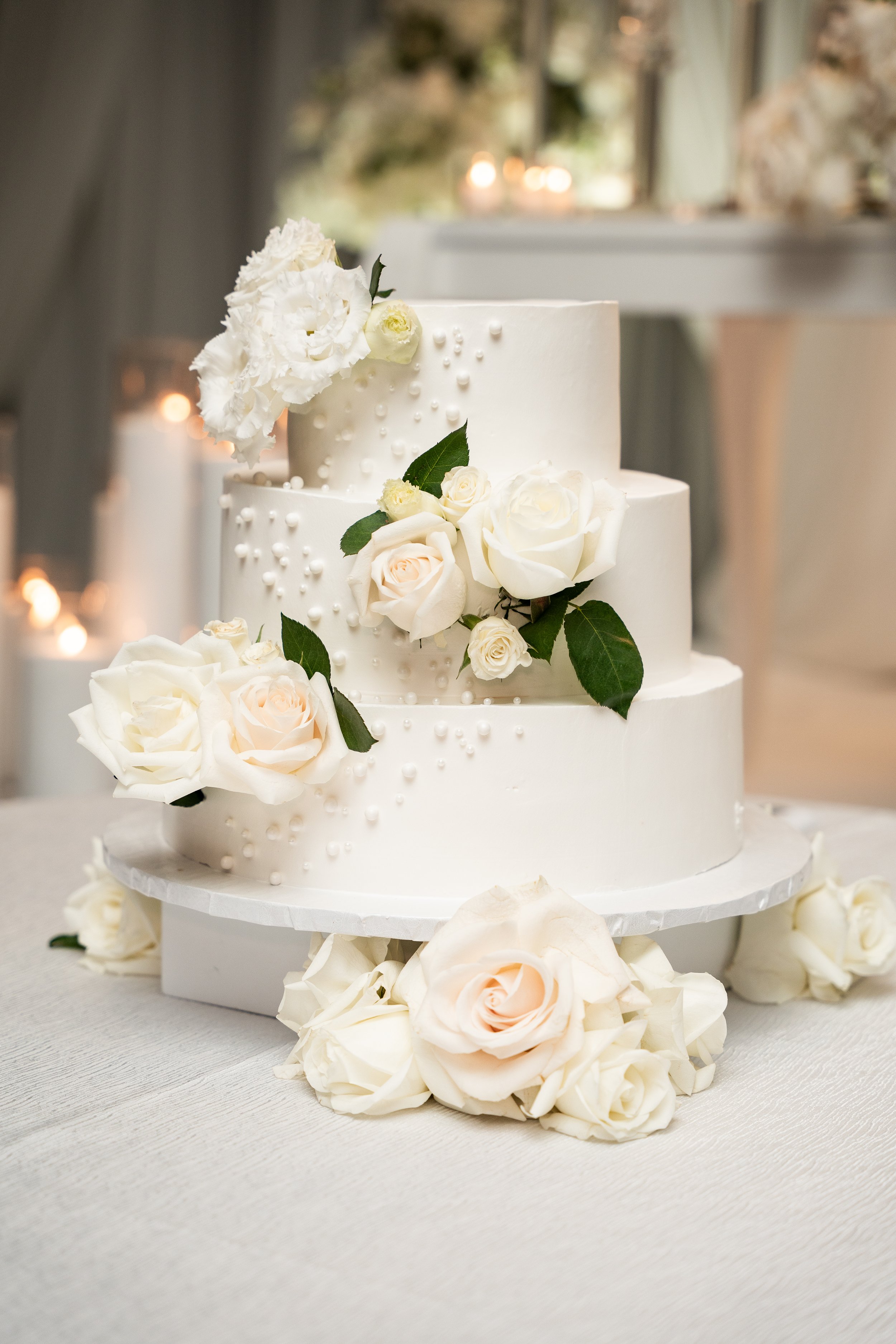 Two-tiered white wedding cake decorated with white roses, light pink roses, and green leaves, with delicate white pearl details, placed on a white cake stand surrounded by white rose petals.