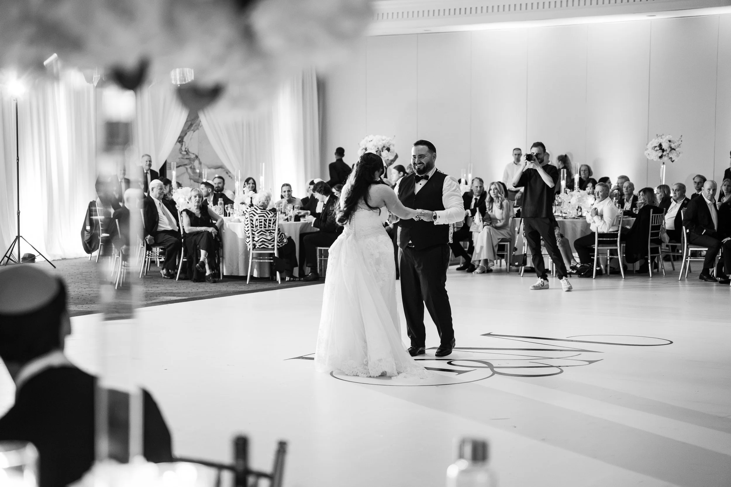A bride and groom dancing at their wedding reception. The bride is in a white gown and the groom is in a black vest and bow tie. Guests are seated at decorated tables in the background, with a photographer capturing the moment.