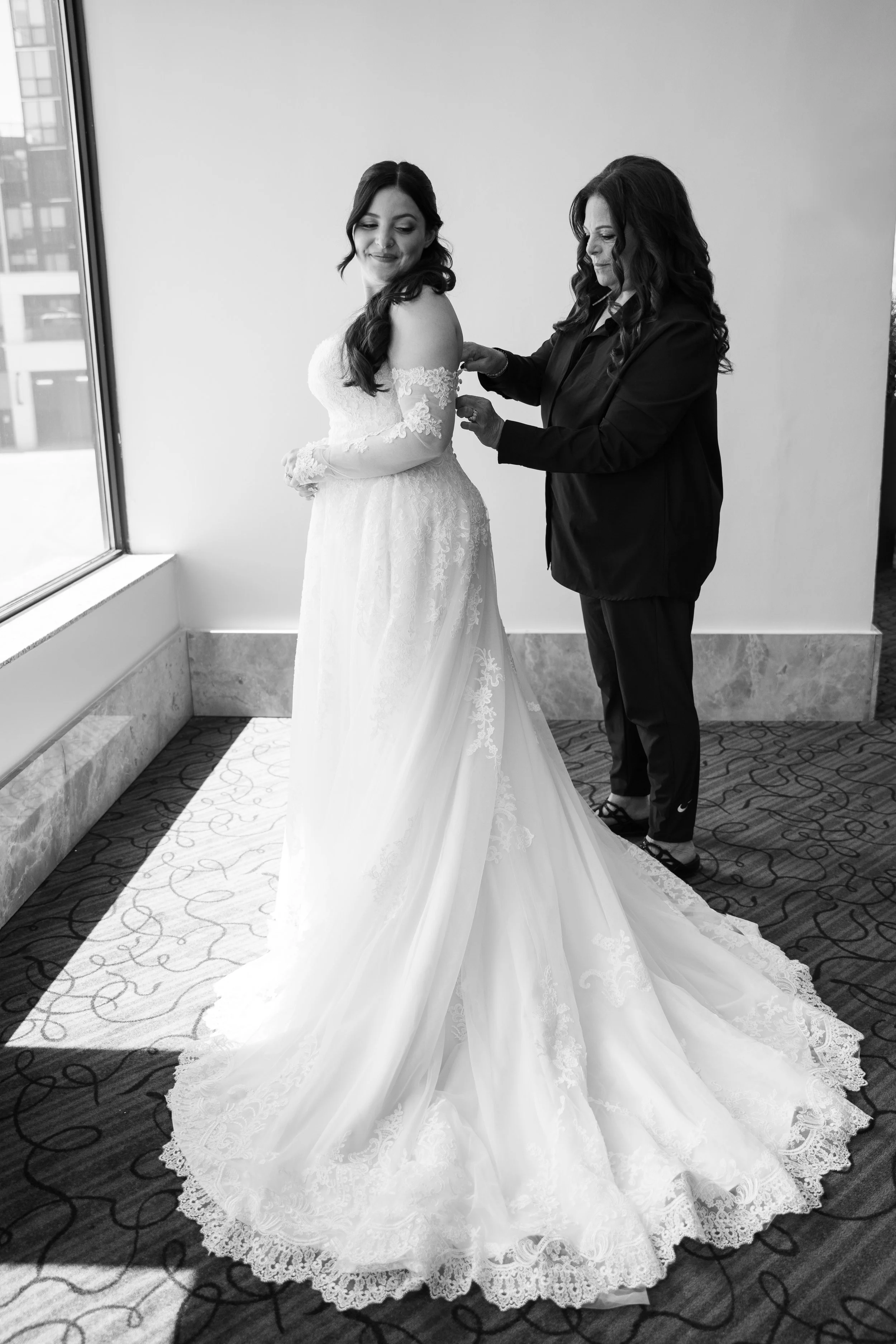 A woman in a wedding dress stands near a window, while another woman helps her adjust her dress. The wedding dress features lace details with a flowing train, and the scene is indoors near a patterned carpet.
