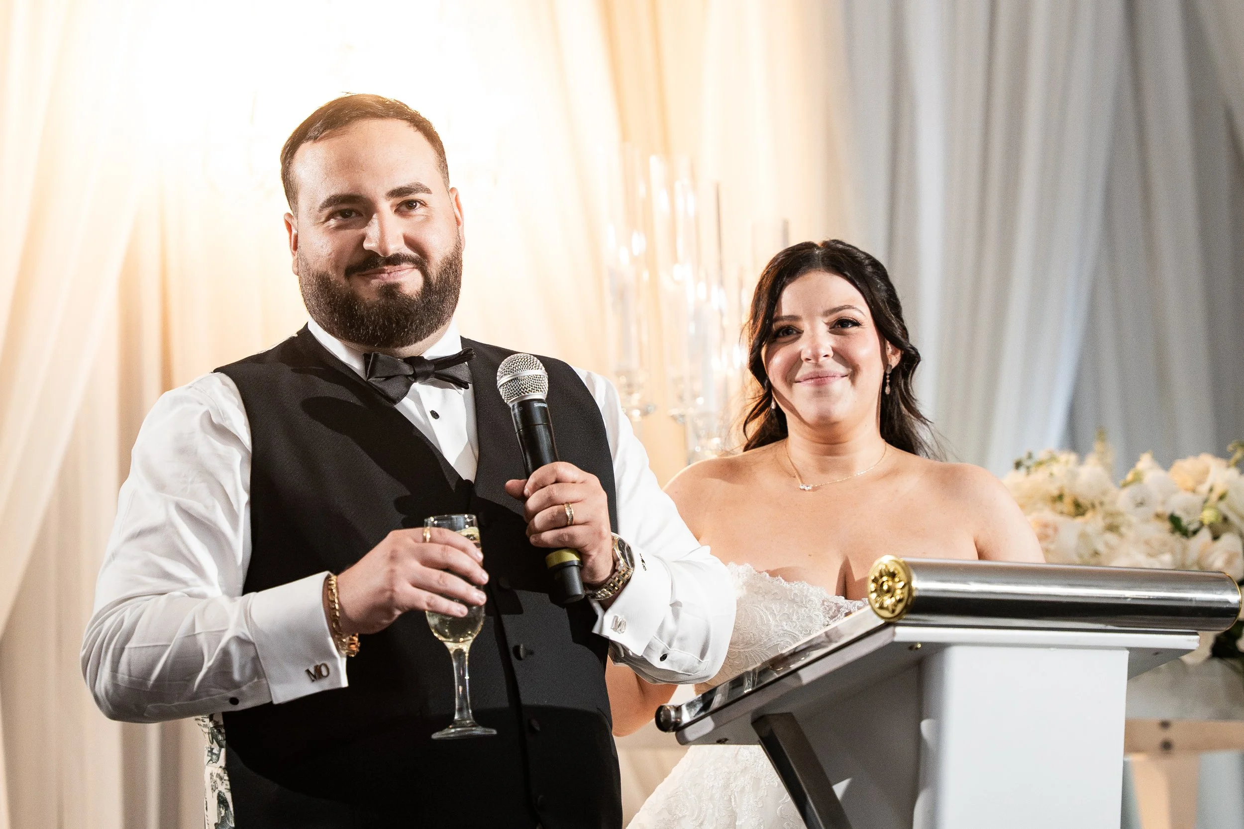 Man in tuxedo giving a speech at a wedding reception, holding a microphone, with a woman in a wedding dress standing beside him, in front of floral arrangements and curtains.
