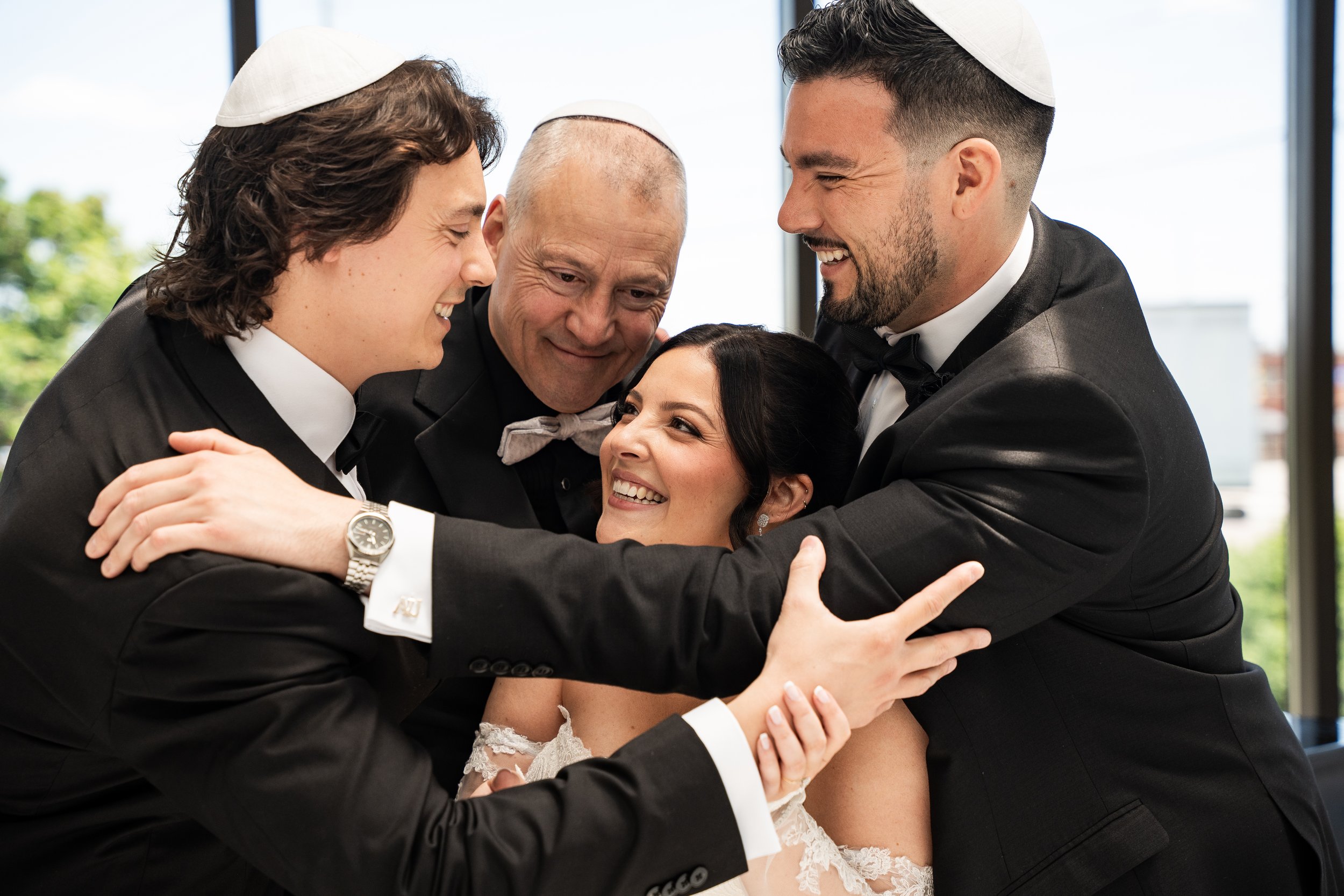 A group of five people, including a bride and four men, are smiling and hugging each other joyfully during a celebration. All are wearing formal Black tie attire and yarmulkes.