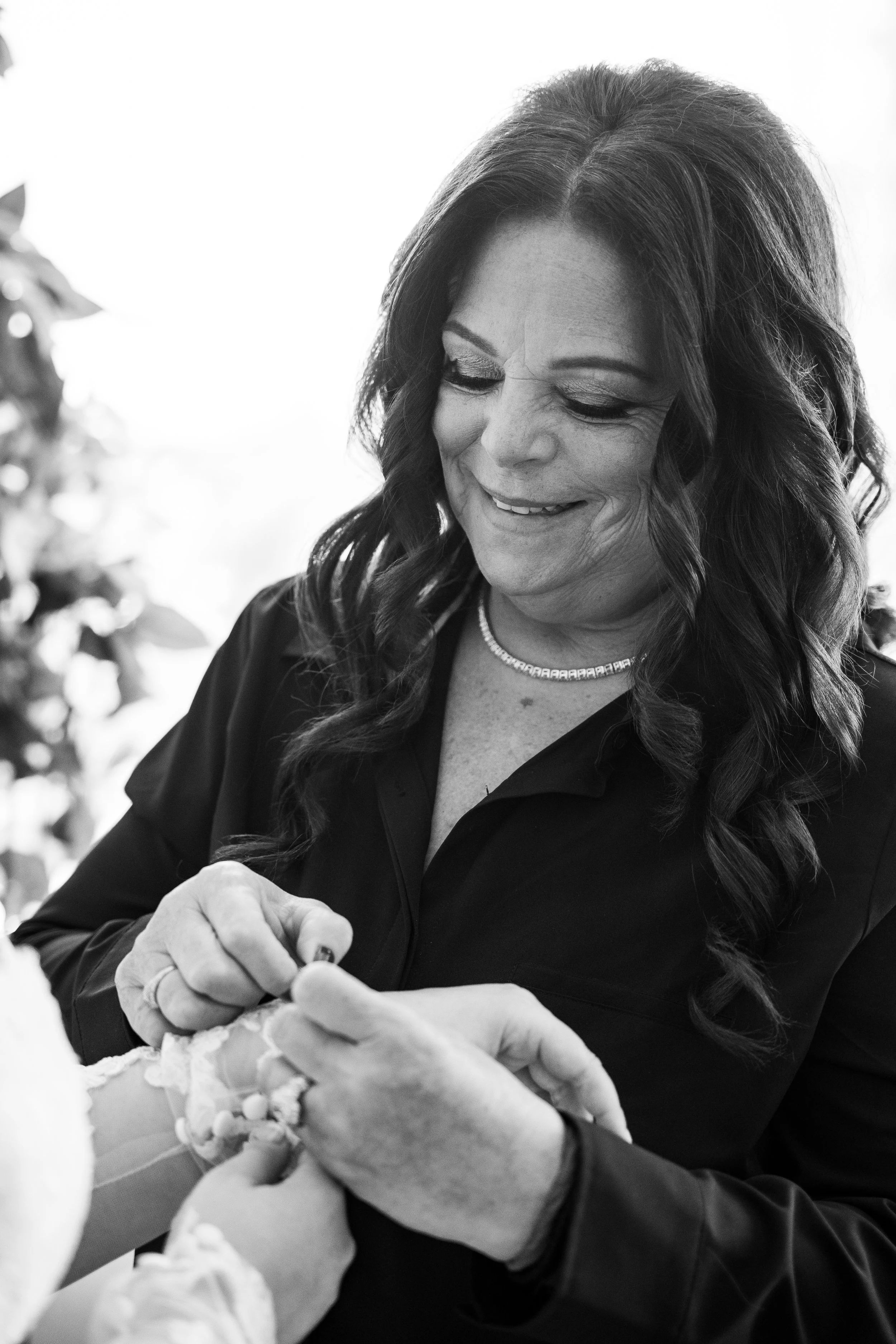 A woman with long, wavy hair, wearing a dark shirt and a necklace, smiles while receiving a manicure from another person.