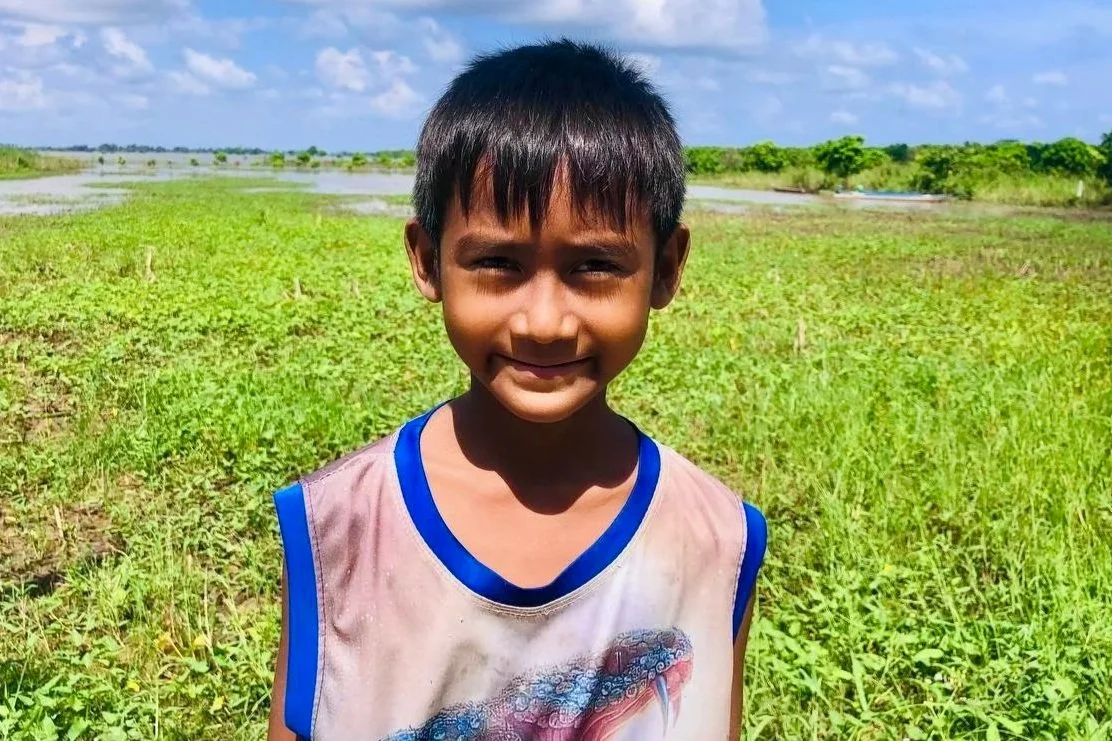 Smiling boy standing in a lush green field under a bright blue sky with fluffy clouds, conveying a cheerful and serene countryside vibe.