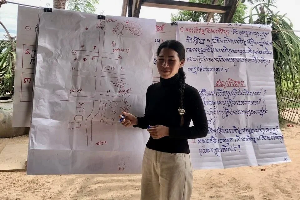 A woman in a black top and light pants stands in front of large handwritten papers with a map and text. She is outdoors, near palm trees, appearing focused.