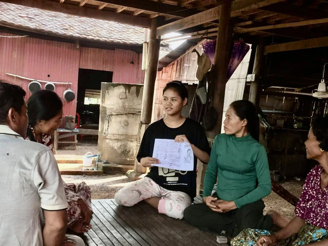 A young woman holds educational materials, engaged in discussion with four attentive adults seated around her in a rustic, wooden interior setting.