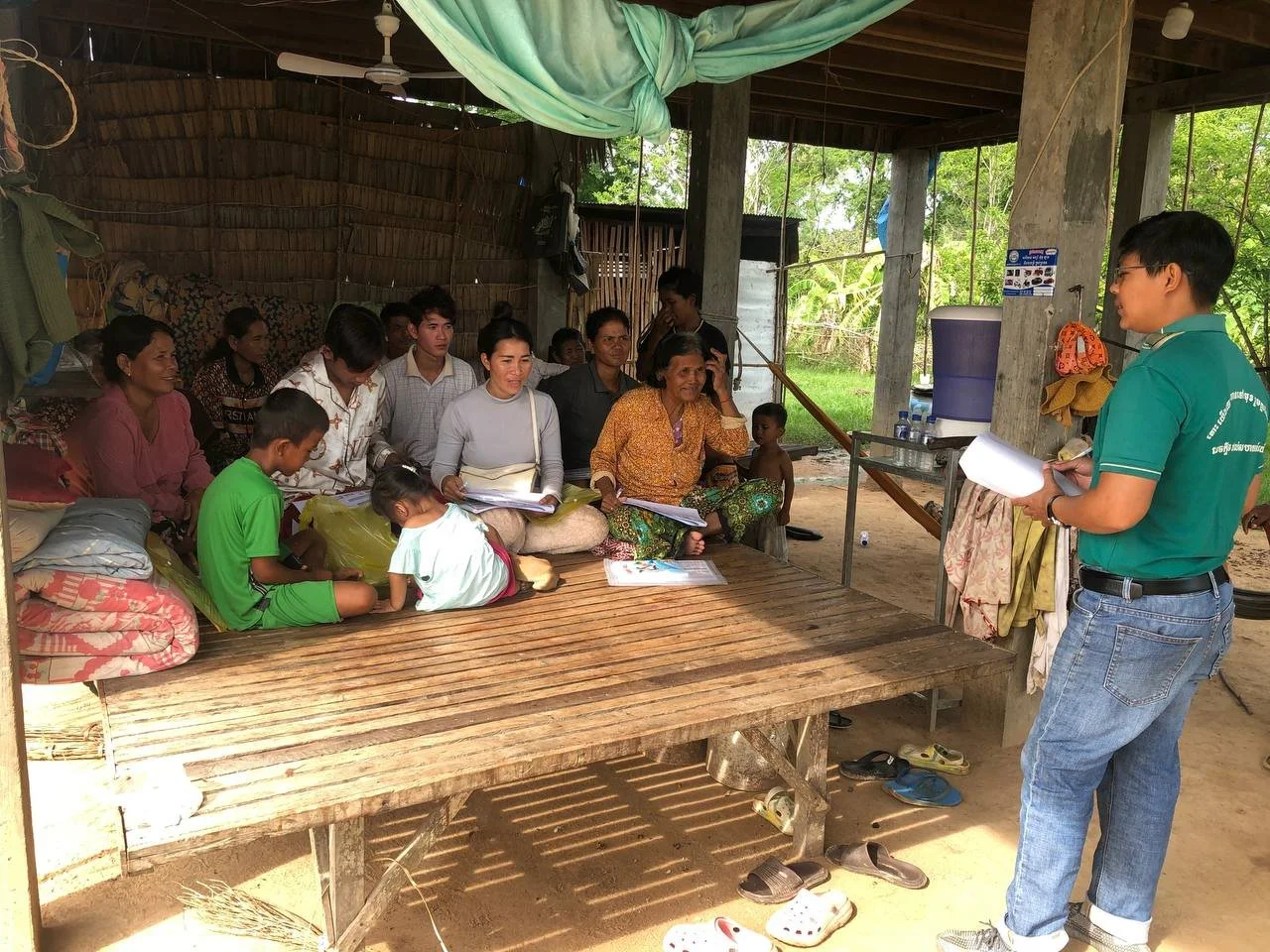 A community meeting takes place under an open shelter with wooden walls. A group listens intently to a man in a green shirt holding papers. The atmosphere is attentive and engaged.