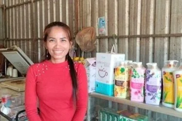 A woman in a red sweater smiles warmly behind a store counter displaying colorful bottles of products. The setting has a cozy, rustic feel.