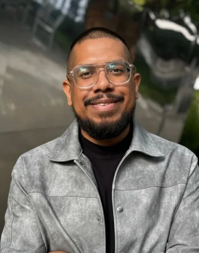 Smiling man with glasses, short hair, and a beard, wearing a black shirt, against a plain white background.