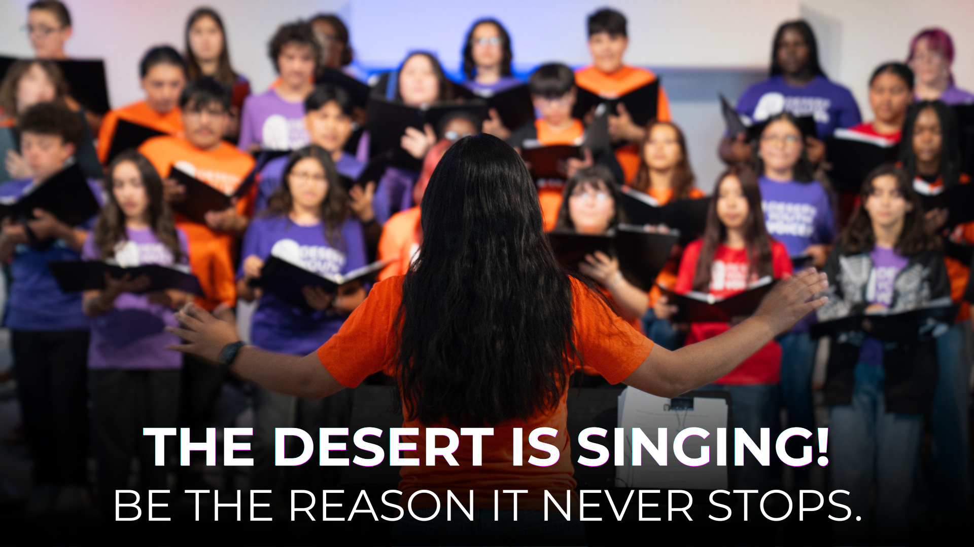 A choir of children singing, led by a woman with long black hair and an orange shirt, in front of a plain white wall.