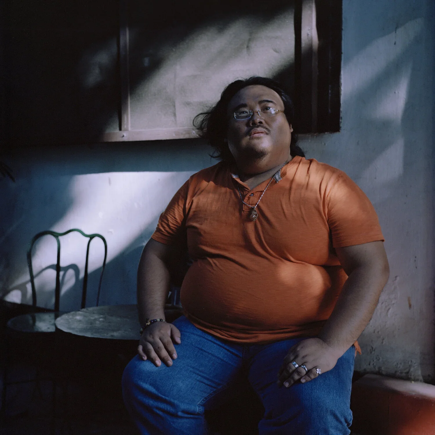 Ein Mann sitzt in einem Raum mit dunklem Hintergrund, trägt eine orange Hemd und blaue Jeans, und blickt nach oben.