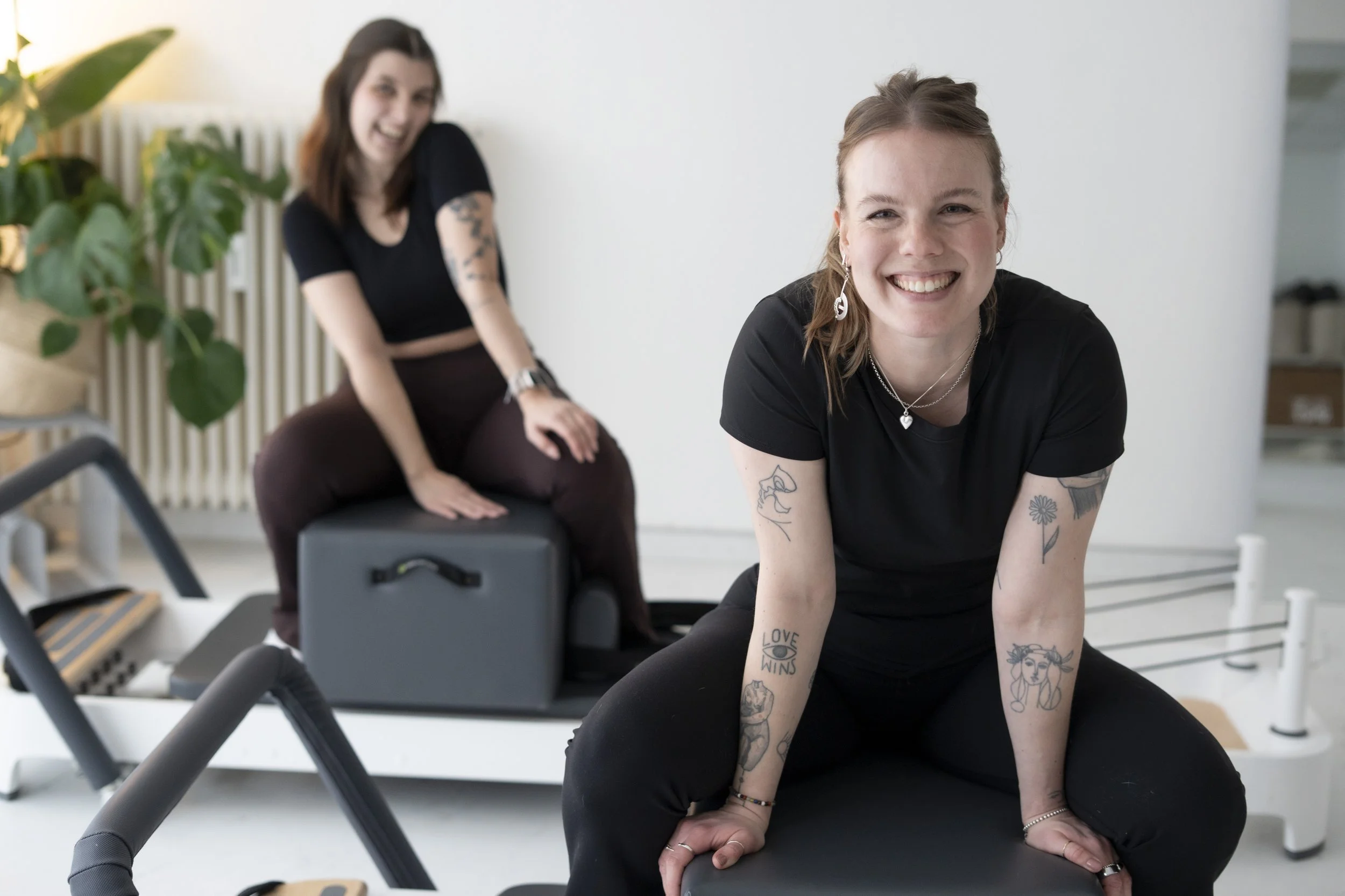 Zwei lachende Frauen, eine sitzt auf einem Fitnessgerät, die andere kniet vor ihr. Das Bild ist in einem hellen Raum mit Pflanzen im Hintergrund.