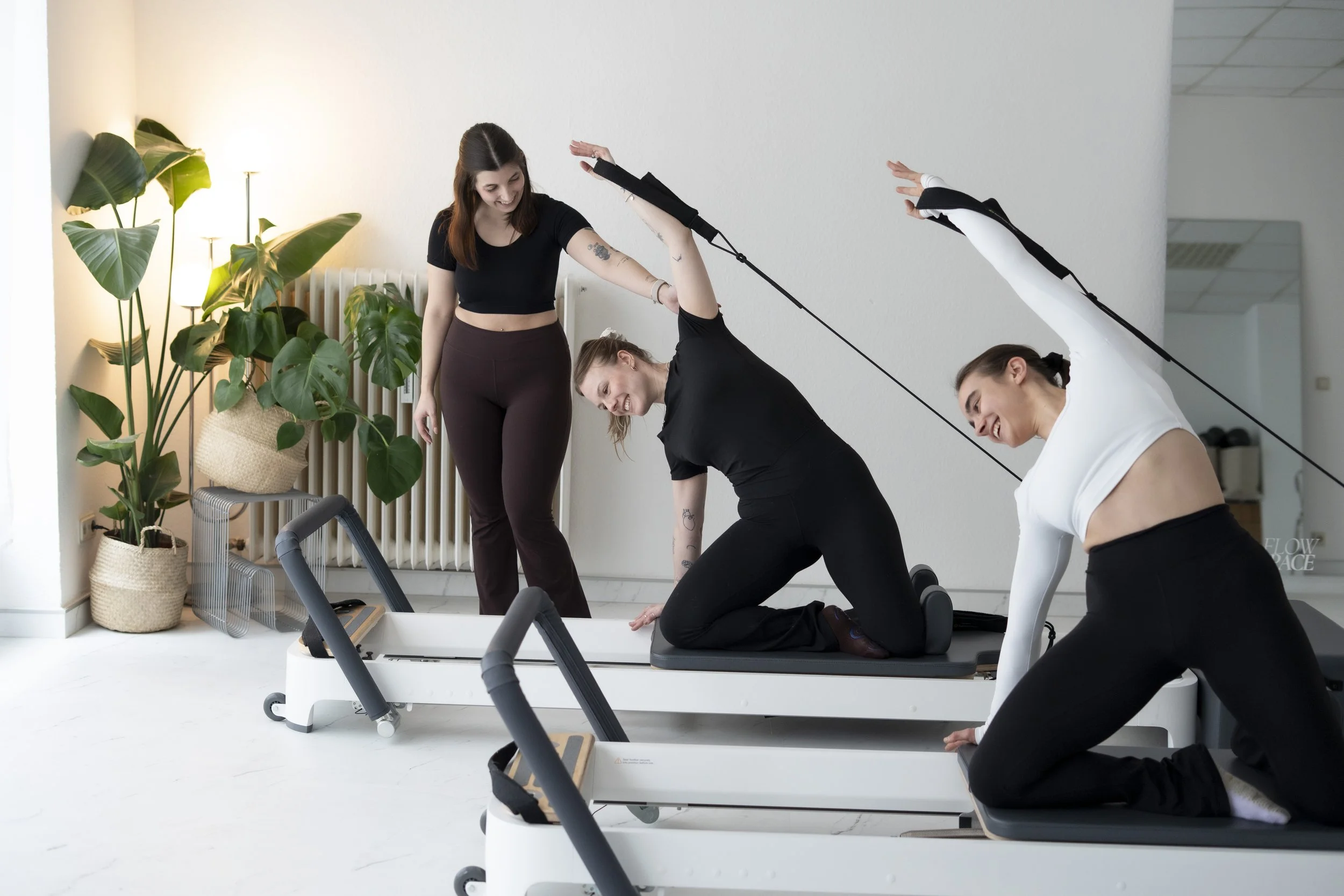 Drei Frauen trainieren im Fitnessstudio auf einem Laufband, eine Frau befindet sich in der Hundestellung, während eine andere Frau die Übungen beobachtet und die dritte Frau in der Nähe bei den Übungen assistiert, in einem hellen Raum mit Pflanzen.