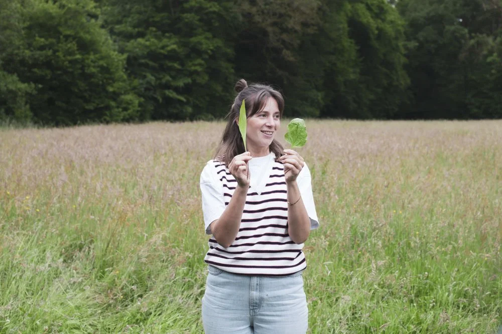 Junge Frau hält zwei grüne Blätter, eines in jeder Hand, im Freien auf einer Wiese mit Bäumen im Hintergrund. Pia von Krautfounding, Köln.
