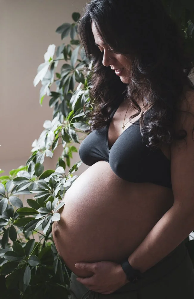 Schwangere Frau in schwarzer Sport-BH, die ihren Babybauch vor grünen Pflanzen hält, lächelnd, bei natürlichem Licht. Babybauch Shooting, Schmelz.