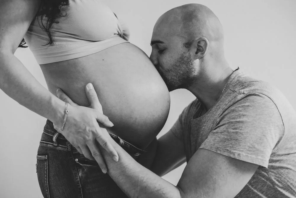 Ein Mann küsst den Babybauch einer schwangeren Frau, beide in einem Schwarz-Weiß-Bild, im intimvollen Moment. Babybauchfotografie.