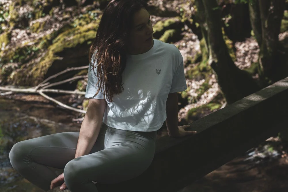 Frau sitzt auf einem Baumstamm in einem Wald, trägt weiße Kleidung und hat braune, lockige Haare.