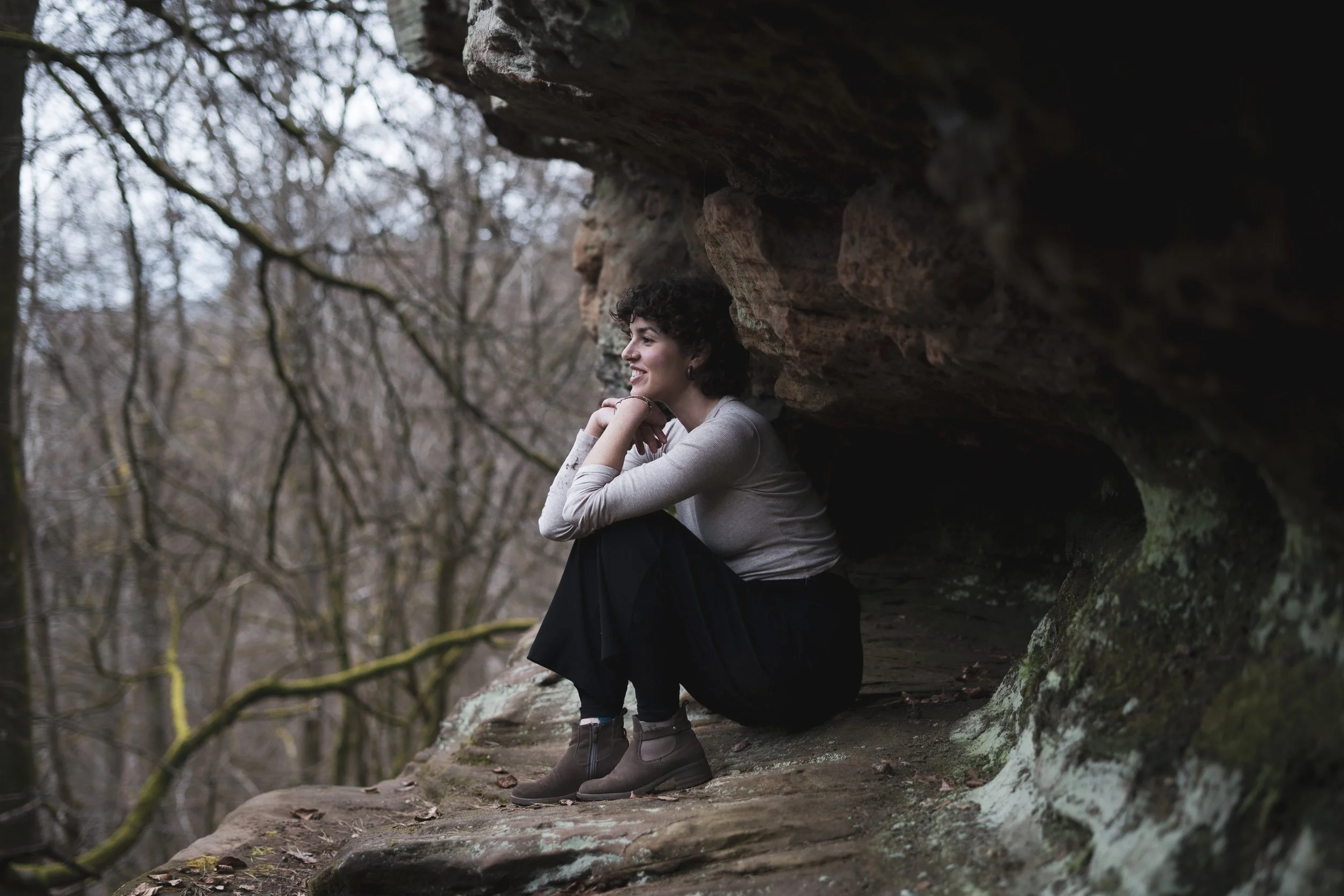 Eine lachende Frau sitzt auf einem Felsen im Wald, trägt eine graue Strickjacke, schwarze Hose und braune Stiefel, im Hintergrund sind Bäume ohne Blätter.