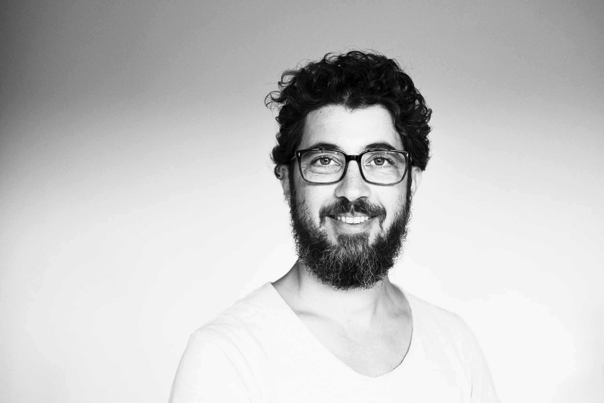Schwarz-Weiß-Foto eines lachenden Mannes mit Brille, Bart und lockigem Haar, trägt ein weißes T-Shirt.