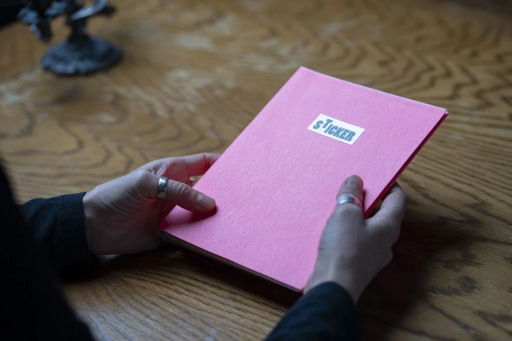 Person hält ein rotes Heft mit der Aufschrift 'Sticker' auf einem Holztisch.