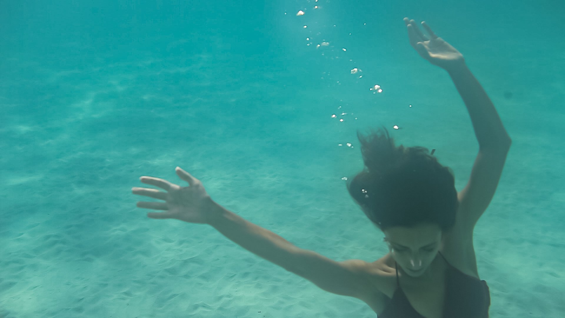Eine Person schwimmt unter Wasser, mit geradem Gesicht nach unten, Arme ausgestreckt, und trägt Sonnenbrille. Die Wasseroberfläche ist sichtbar, Blasen steigen auf.