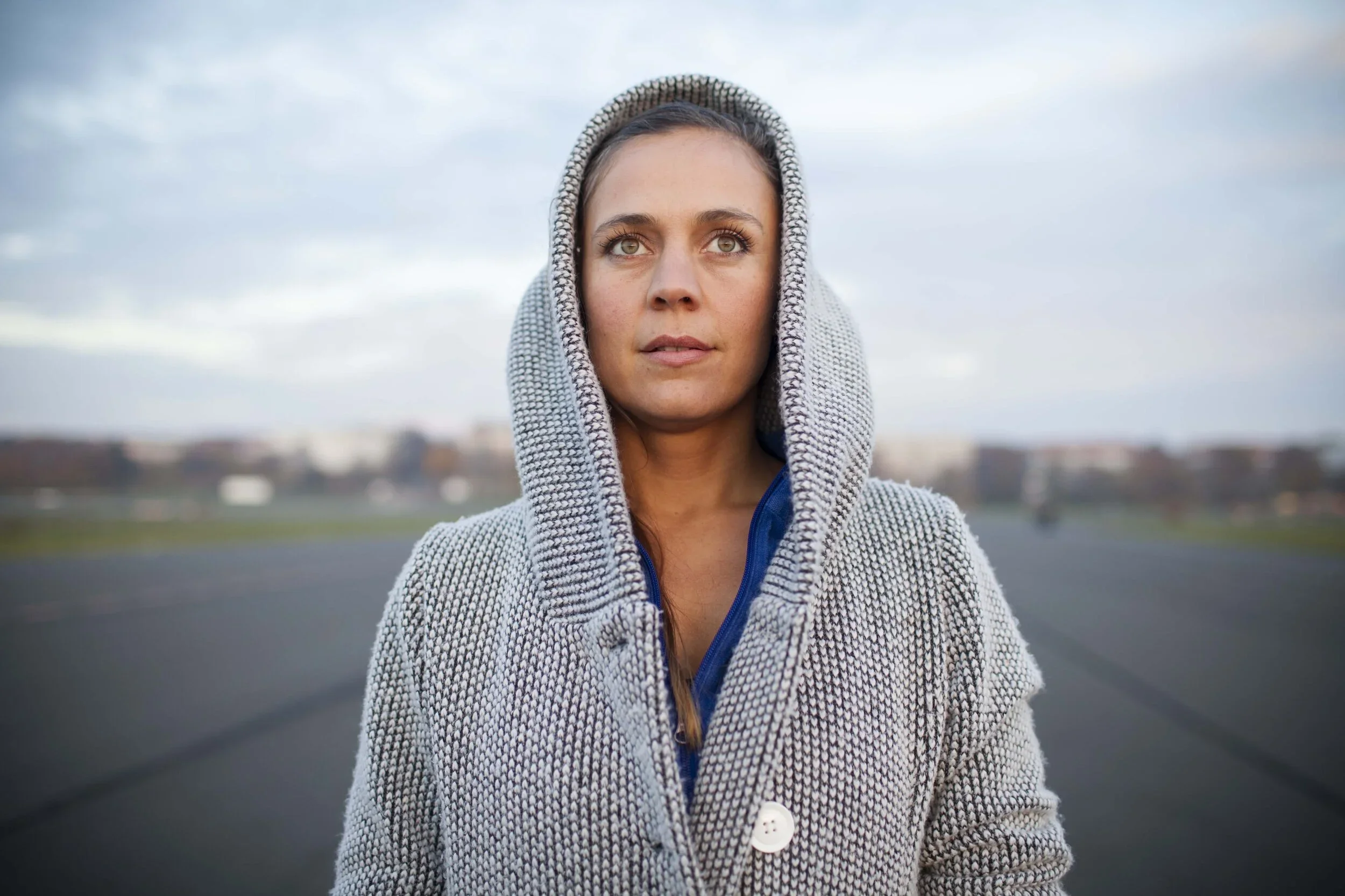 Junge Frau trägt einen gray-blauen Hoodie mit Reißverschluss, blickt nach oben, im Hintergrund eine Straße und ein bewölkter Himmel.