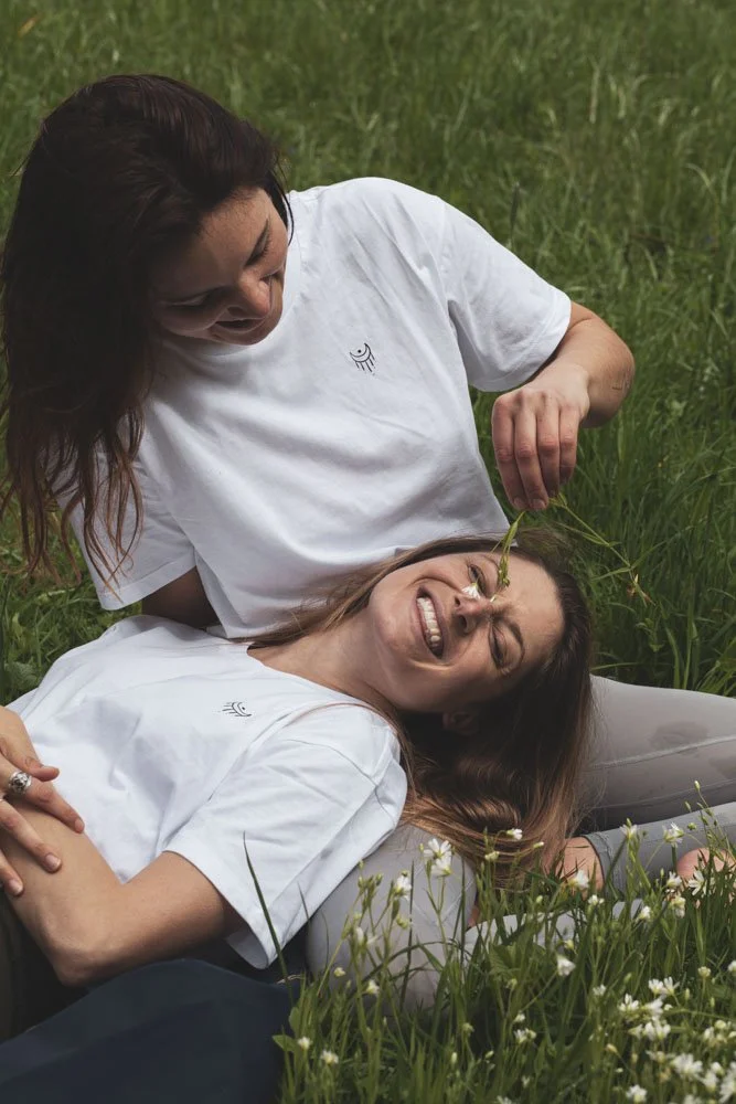 Zwei lachende Frauen liegen im Gras, eine der Frauen hält eine Blume an die Nase der anderen, die sich freut.