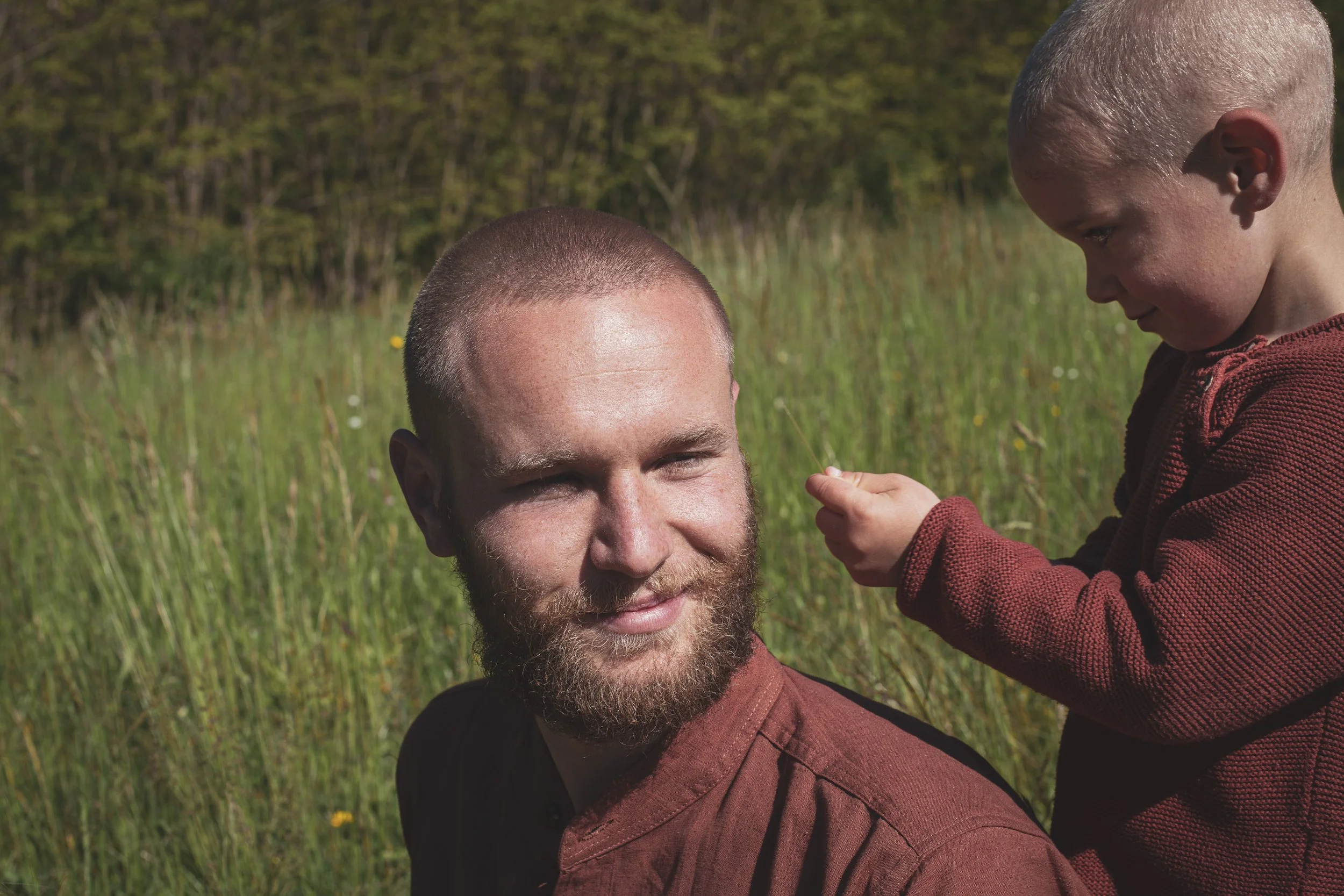 Ein Mann mit Bart und kurzen Haaren sitzt im Freien in einem Feld, während ein kleiner Junge, vermutlich sein Sohn, auf seinem Kopf sitzt und ihn anschaut.