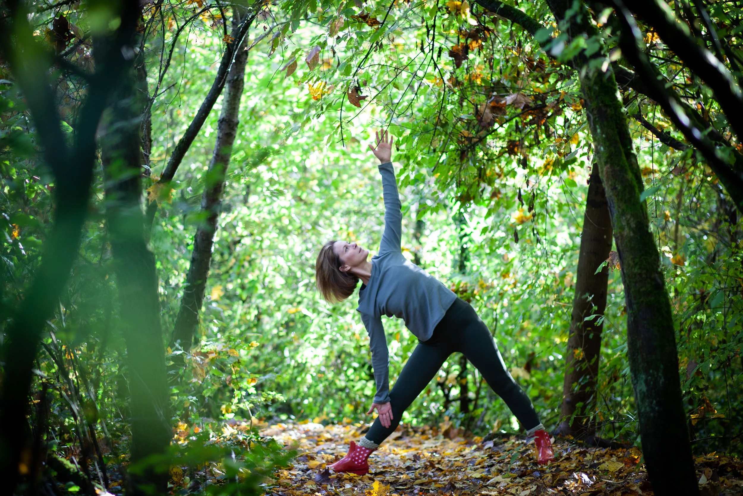 Frau in einem Wald übt Yoga, hält eine Handfläche zur Decke, umgeben von Bäumen und Herbstlaub.