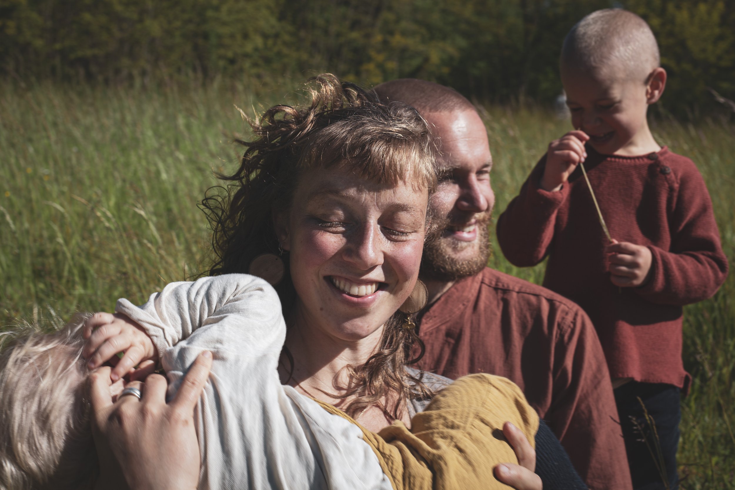 Vier Menschen, zwei Frauen, ein Mann und ein Kind, lachen und umarmen sich in einem grünen Feld bei Sonnenschein.