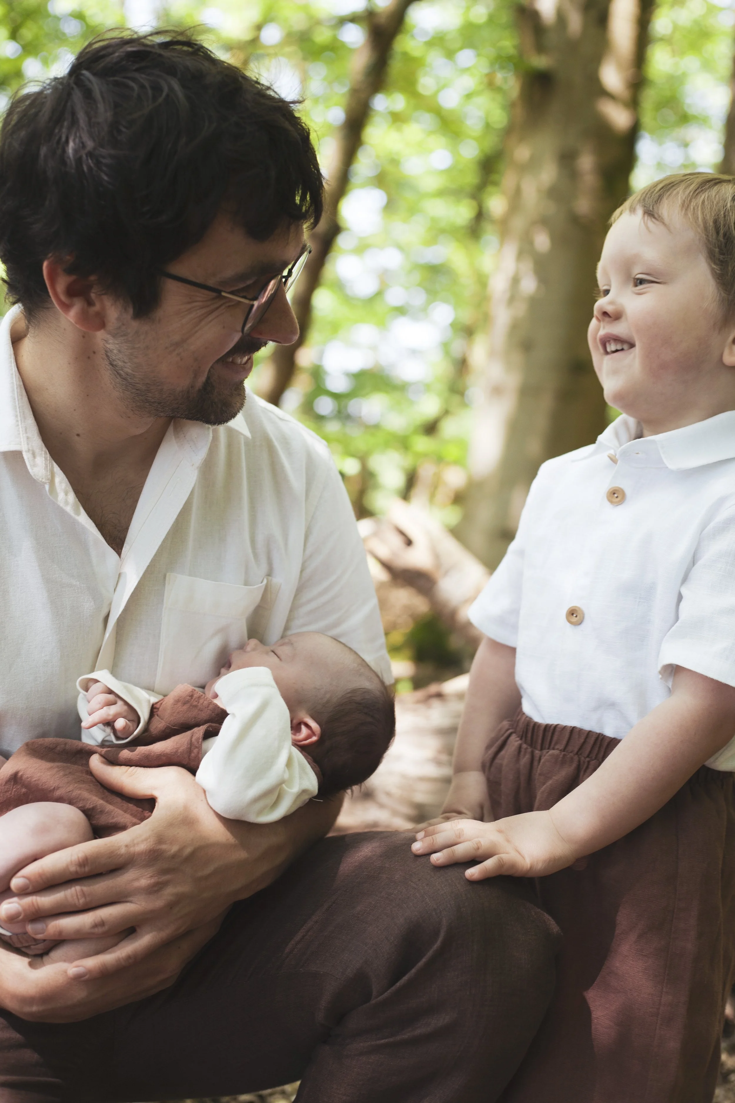 Ein Mann hält ein neugeborenes Baby im Arm, während ein Junge, vermutlich das Geschwisterkind, lachend zugeschaut. Die Szene findet im Freien statt, umgeben von Bäumen und grünem Laub.