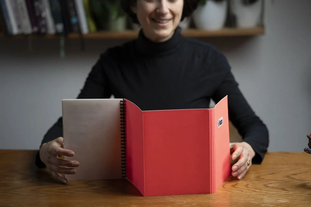 Eine Frau in schwarzem Rollkragenpullover sitzt an einem Holztisch und hält ein offenes, rotes Buch, das sie betrachtet. Im Hintergrund sind Bücherregale und Pflanzen sichtbar.