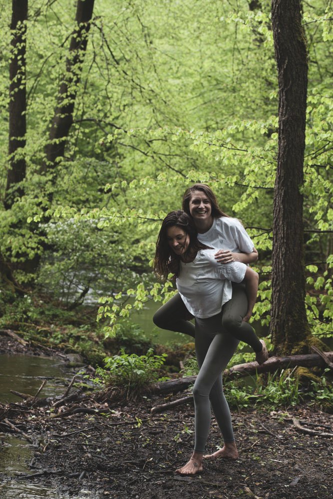 Zwei lachende Frauen im Wald, eine trägt die andere auf ihrem Rücken, barfuß auf matschigem Boden am Ufer eines kleinen Baches, umgeben von grünen Blättern und Bäumen.