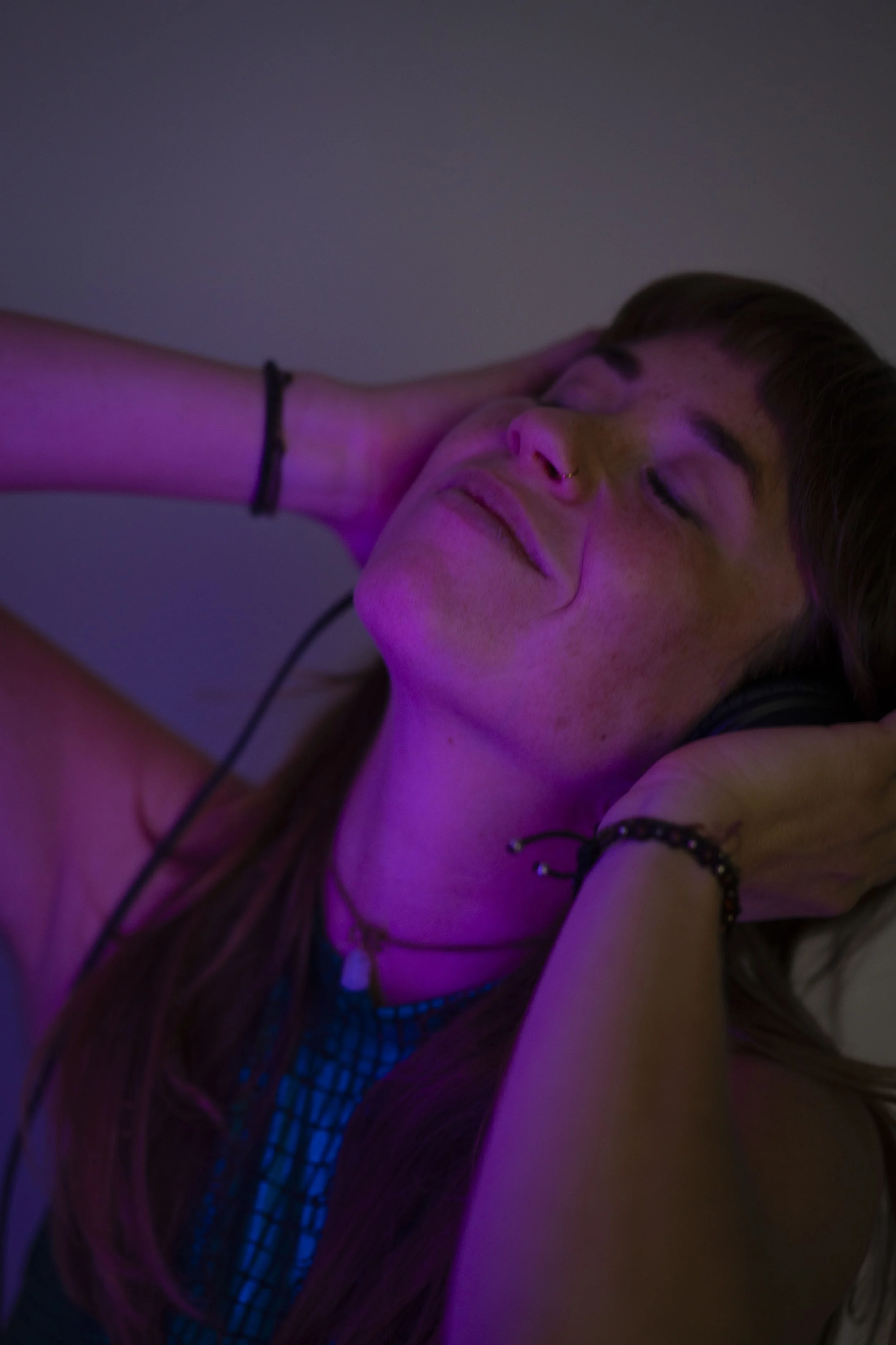 Junge Frau liegt mit geschlossenen Augen, berührt ihren Hals mit der Hand, trägt einen Nasenring, mehrere Armbänder, headphones und ein träumerisches Gesicht.