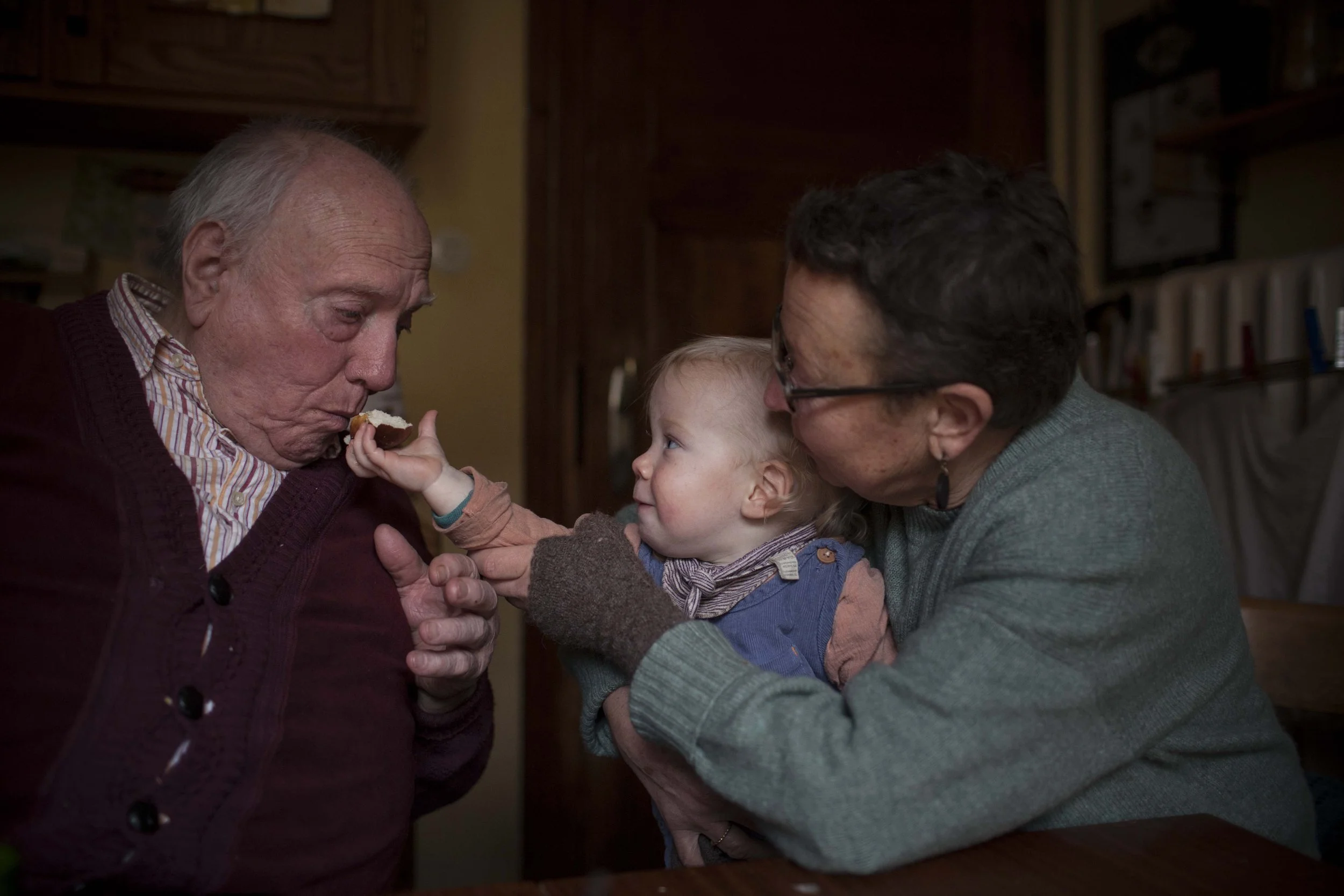 Ein älterer Mann wird von einem kleinen Kind mit einem Stück Kuchen gefüttert, während eine Frau mit kurzen dunklen Haaren und Brille zuschaut. Das Bild zeigt eine familiäre Szene in einem gemütlichen Raum.