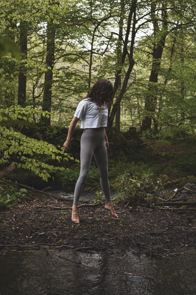 Eine Frau steht barfuß an einem Bach im Wald, trägt ein weißes T-Shirt und graue Leggings, umgeben von grünen Bäumen und Laub.