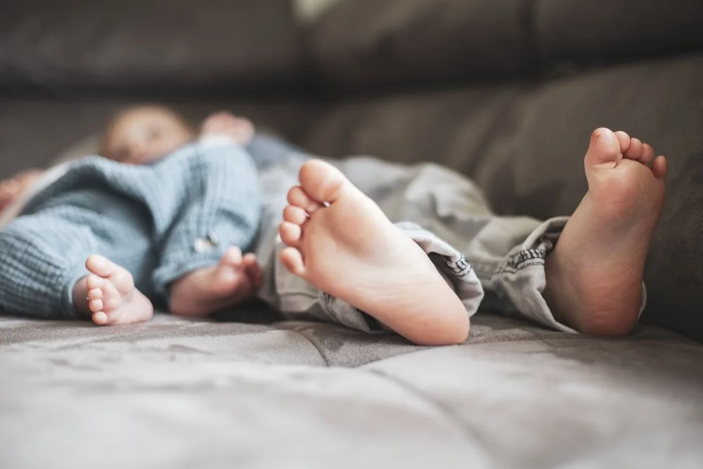 Ein schlafender Junge liegt auf einem Sofa, mit Blick auf seine nackten Füße in der Vordergrund. Newborn Fotografie mit Geschwisterkind. Homburg, Saarland.