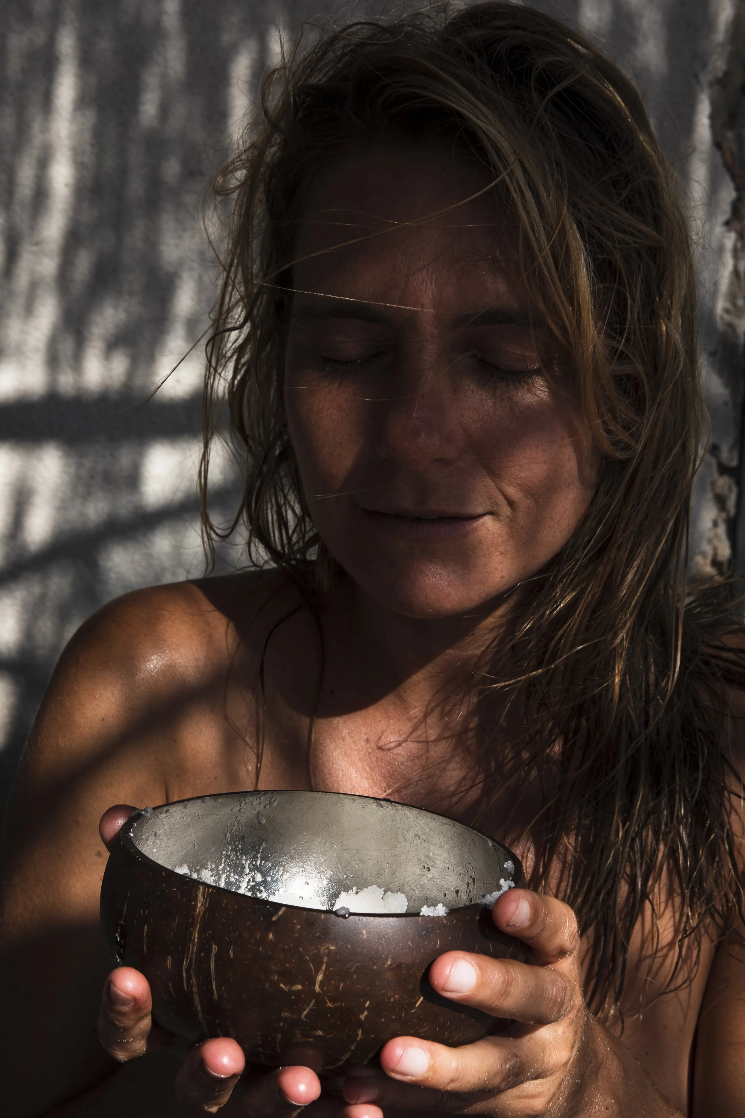 Eine junge Frau mit nassen Haaren hält einen dunklen, runden Becher, aus dem Wasser oder Salz schwappt. Sie ist im Freien, im Sonnenlicht, und hat einen entspannten Ausdruck mit geschlossenen Augen.